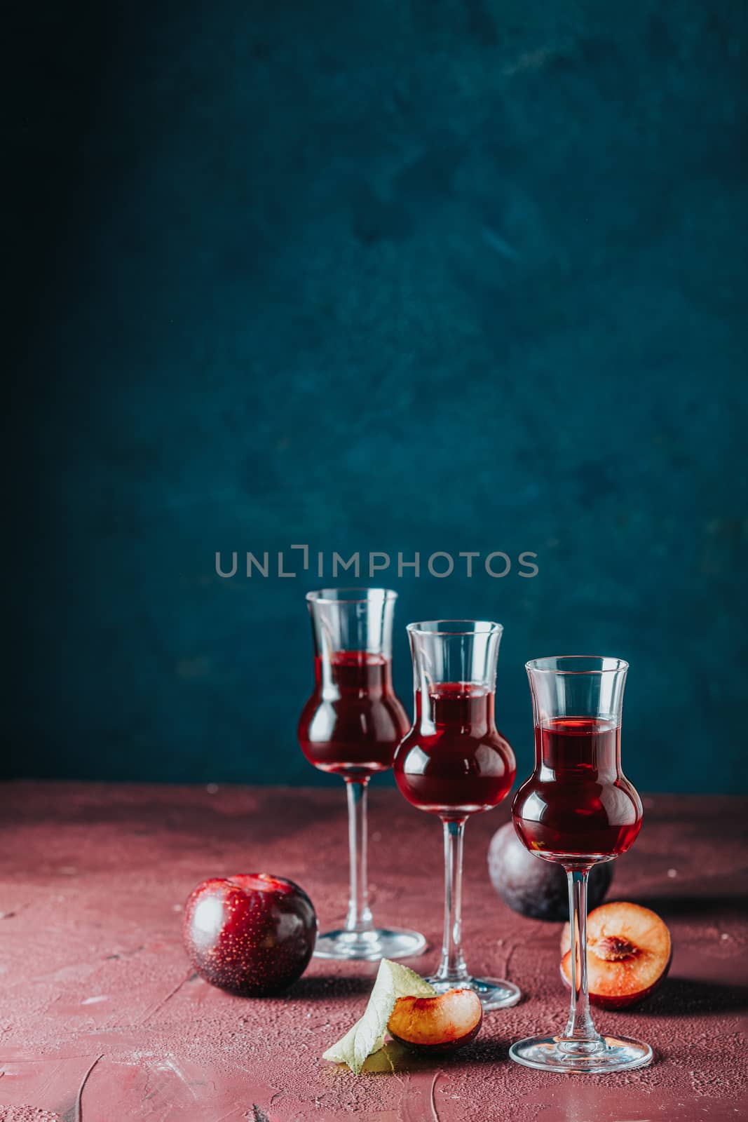 Plums strong alcoholic drink in grappas wineglass with dew. Hard liquor, slivovica, plum brandy or plum vodka with ripe plums on dark blue and claret bordeaux concrete surface