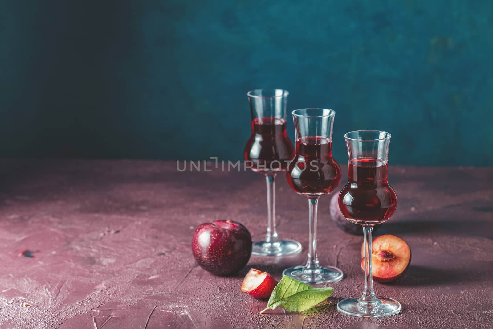 Plums strong alcoholic drink in grappas wineglass with dew. Hard liquor, slivovica, plum brandy or plum vodka with ripe plums on dark blue and  claret bordeaux concrete surface.