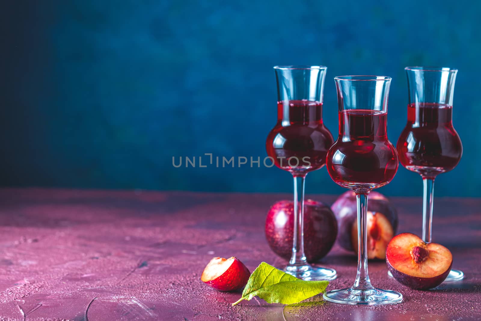 Plums strong alcoholic drink in grappas wineglass with dew. Hard liquor, slivovica, plum brandy or plum vodka with ripe plums on dark blue and claret bordeaux concrete surface