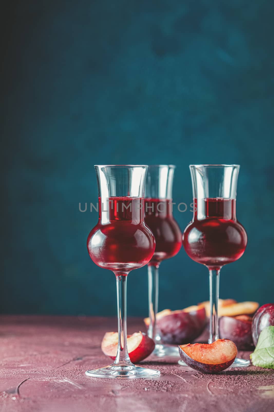 Plums strong alcoholic drink in grappas wineglass with dew. Hard liquor, slivovica, plum brandy or plum vodka with ripe plums on dark blue and  claret bordeaux concrete surface.