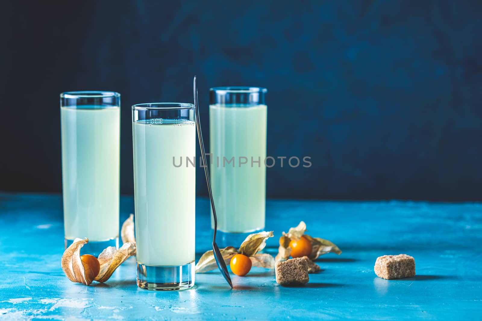 Alcoholic cocktail with Ouzo greek anise brandy or Arabic alcohol drink Raki in highball glass, decorated Physalis peruviana and brown sugar on blue concrete surface.