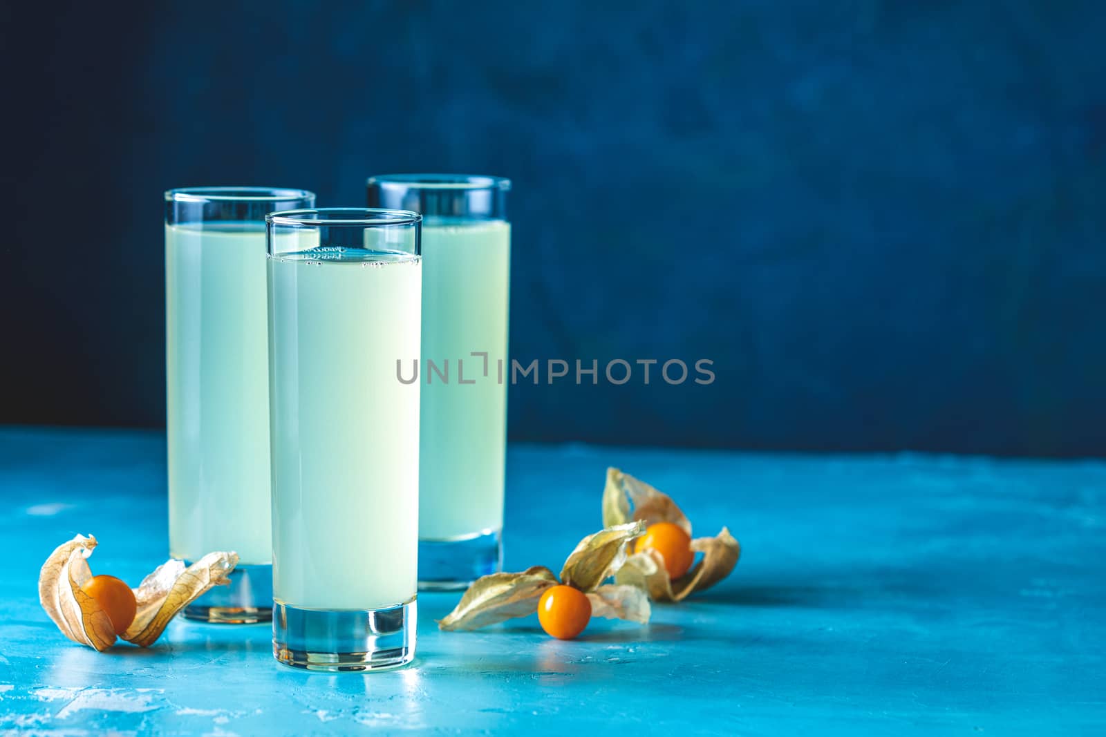 Alcoholic cocktail with Ouzo greek anise brandy or Arabic alcohol drink Raki in highball glass, decorated Physalis peruviana and brown sugar on blue concrete surface.