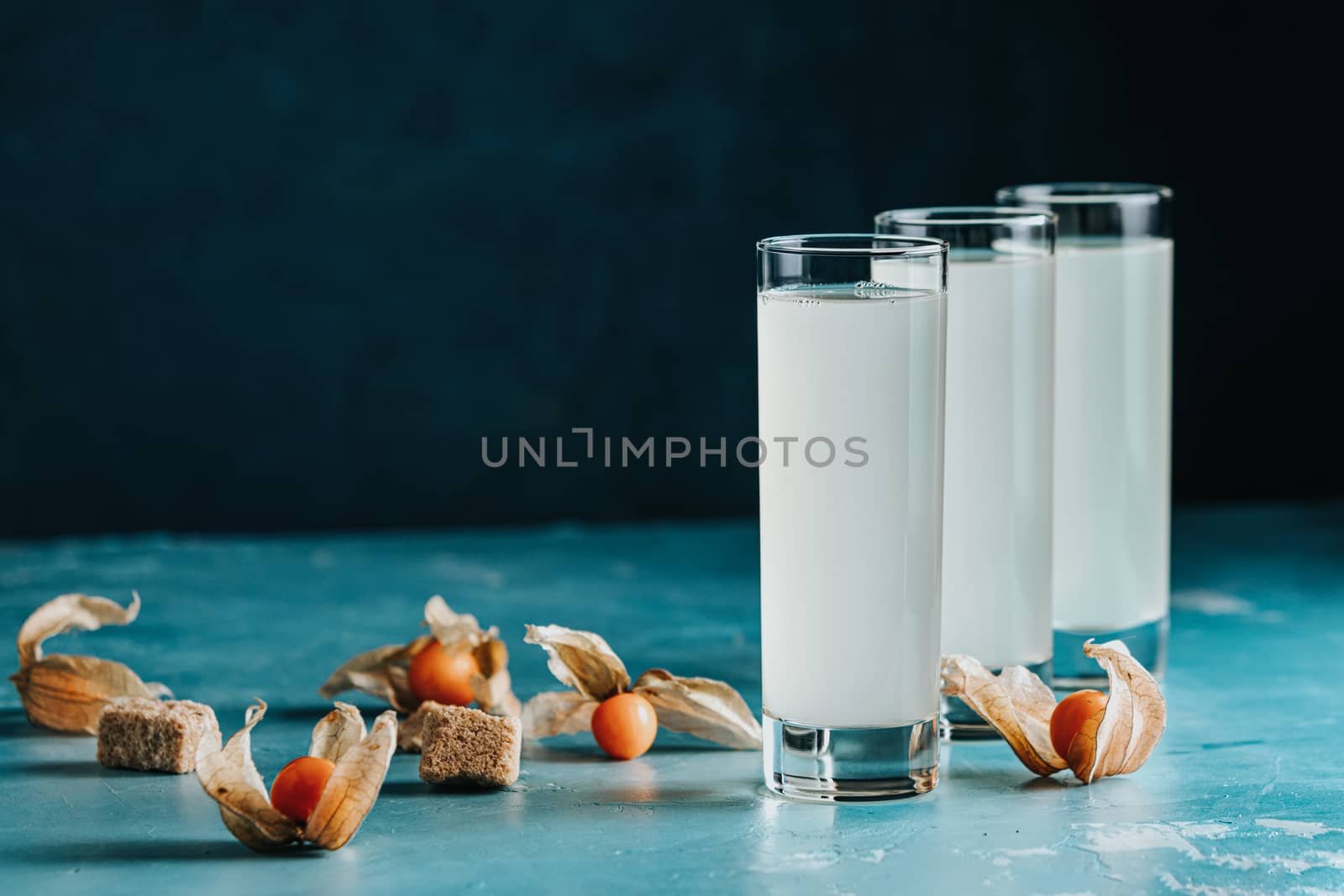 Alcoholic cocktail with Ouzo greek anise brandy or Arabic alcohol drink Raki in highball glass, decorated Physalis peruviana and brown sugar on blue concrete surface.