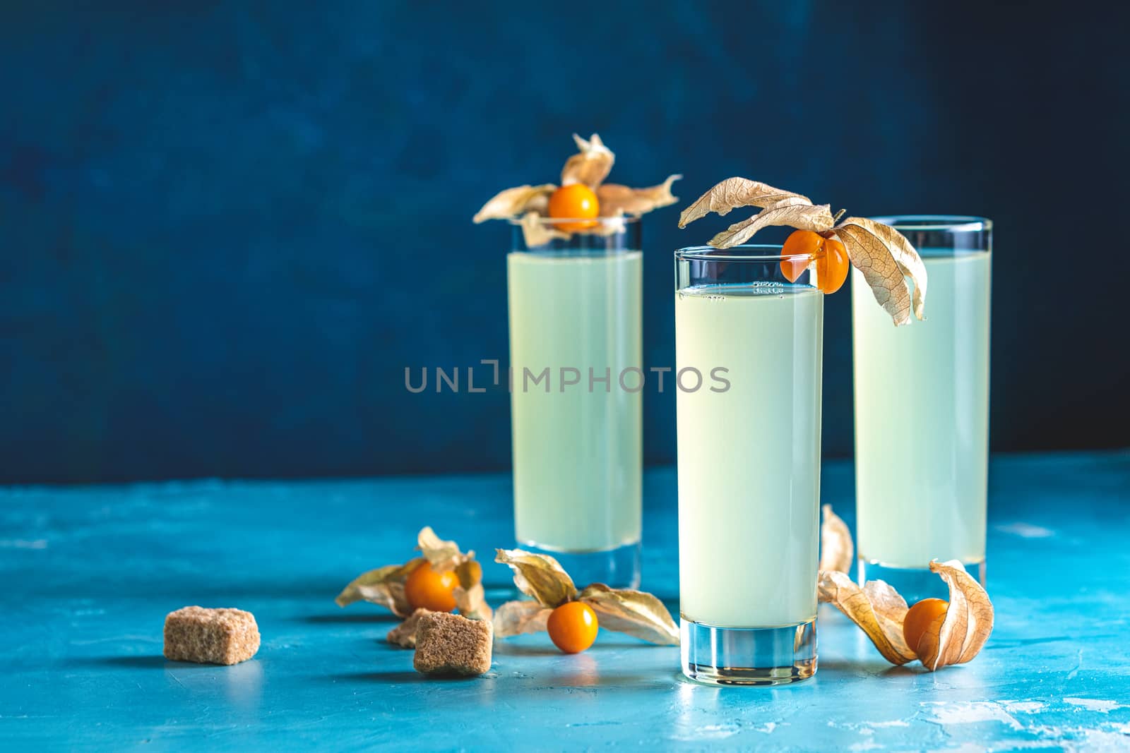 Alcoholic cocktail with Ouzo greek anise brandy or Arabic alcohol drink Raki in highball glass, decorated Physalis peruviana and brown sugar on blue concrete surface.