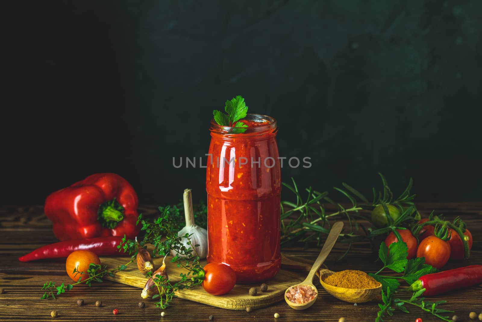 Homemade DIY natural canned hot tomato sauce chutney with chilli or adjika in glass jar standing on wooden table with cherry tomatoes, salt, pepper, herbs, selective focus