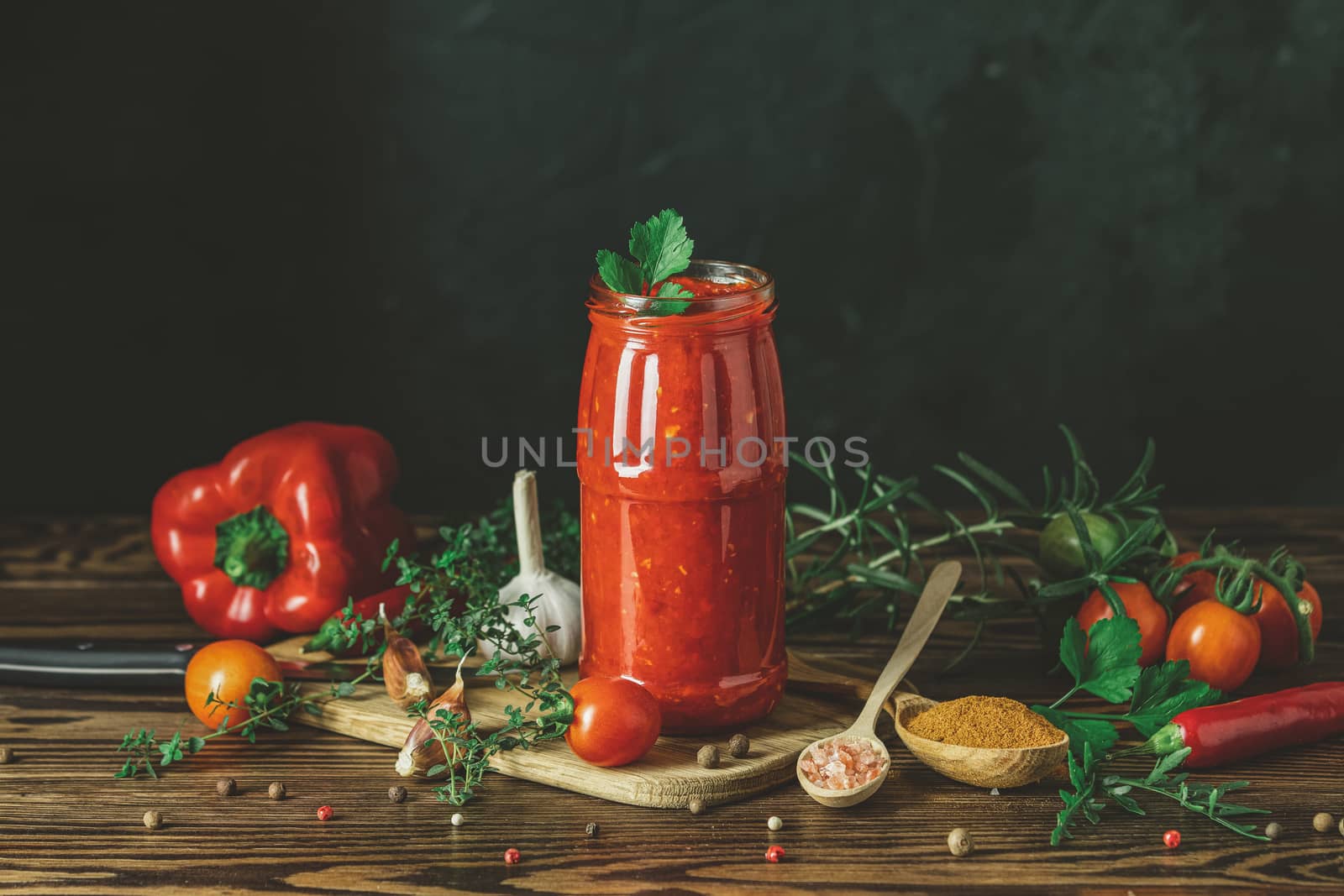 Homemade DIY natural canned hot tomato sauce chutney with chilli or adjika in glass jar standing on wooden table with cherry tomatoes, salt, pepper, herbs, selective focus