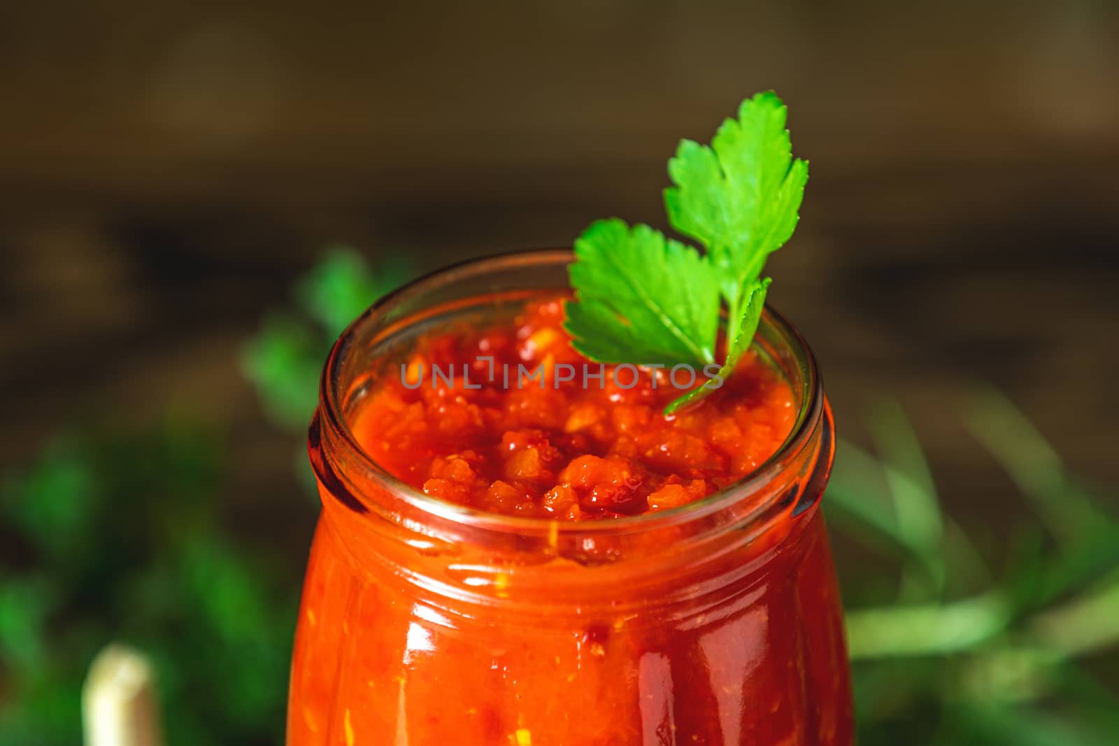 Homemade DIY natural canned hot tomato sauce chutney with chilli or adjika in glass jar standing on wooden table with cherry tomatoes, salt, pepper,  herbs, selective focus