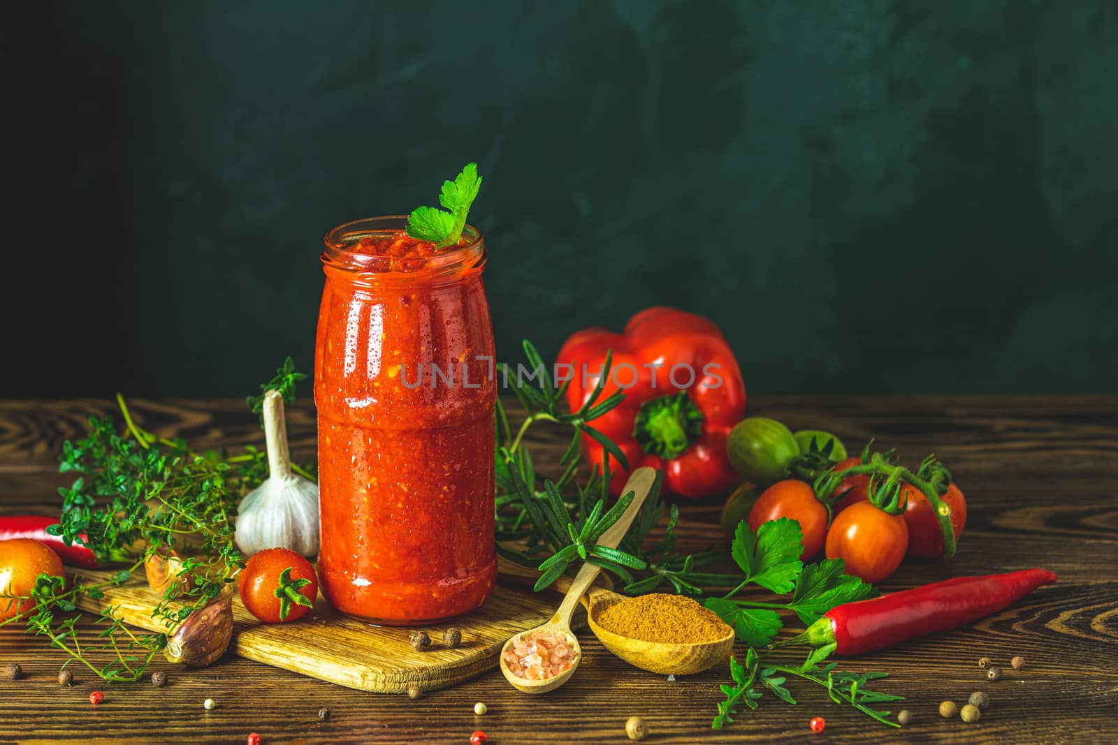 Homemade DIY natural canned hot tomato sauce chutney with chilli or adjika in glass jar standing on wooden table with cherry tomatoes, salt, pepper,  herbs, selective focus