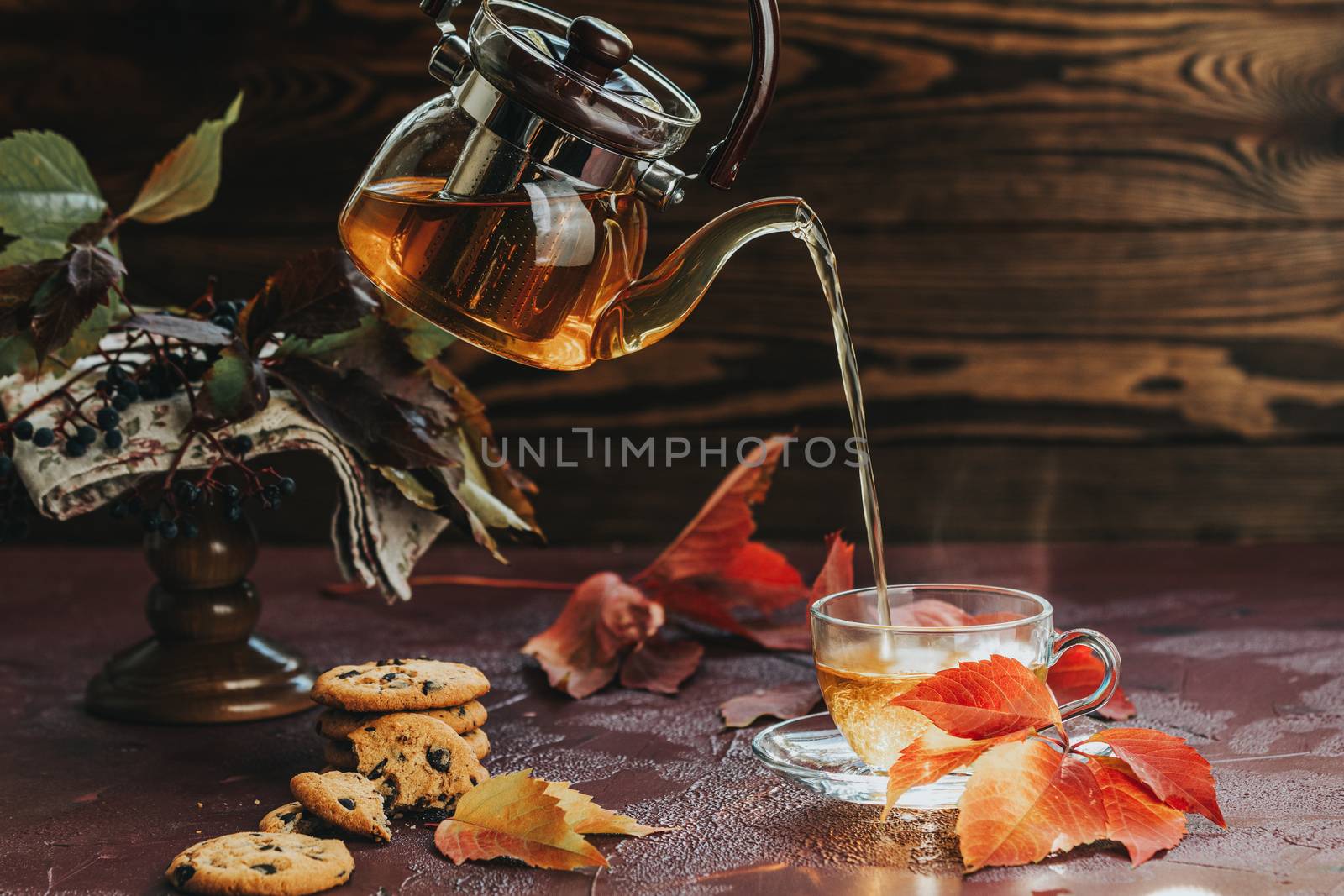 Pour hot tea in glass cup. Autumn teatime composition on dark background with colored leaves and chocolate cookies, sun light beam on the cup.