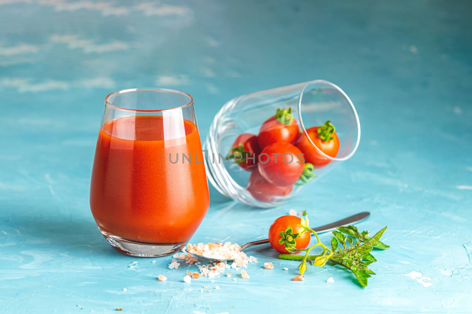 Glasses of fresh delicious jummy red tomato juice and fresh raw tomatoes with pink salt in spoon on light concrete surface. Close up. Gmo free. Natural good food.