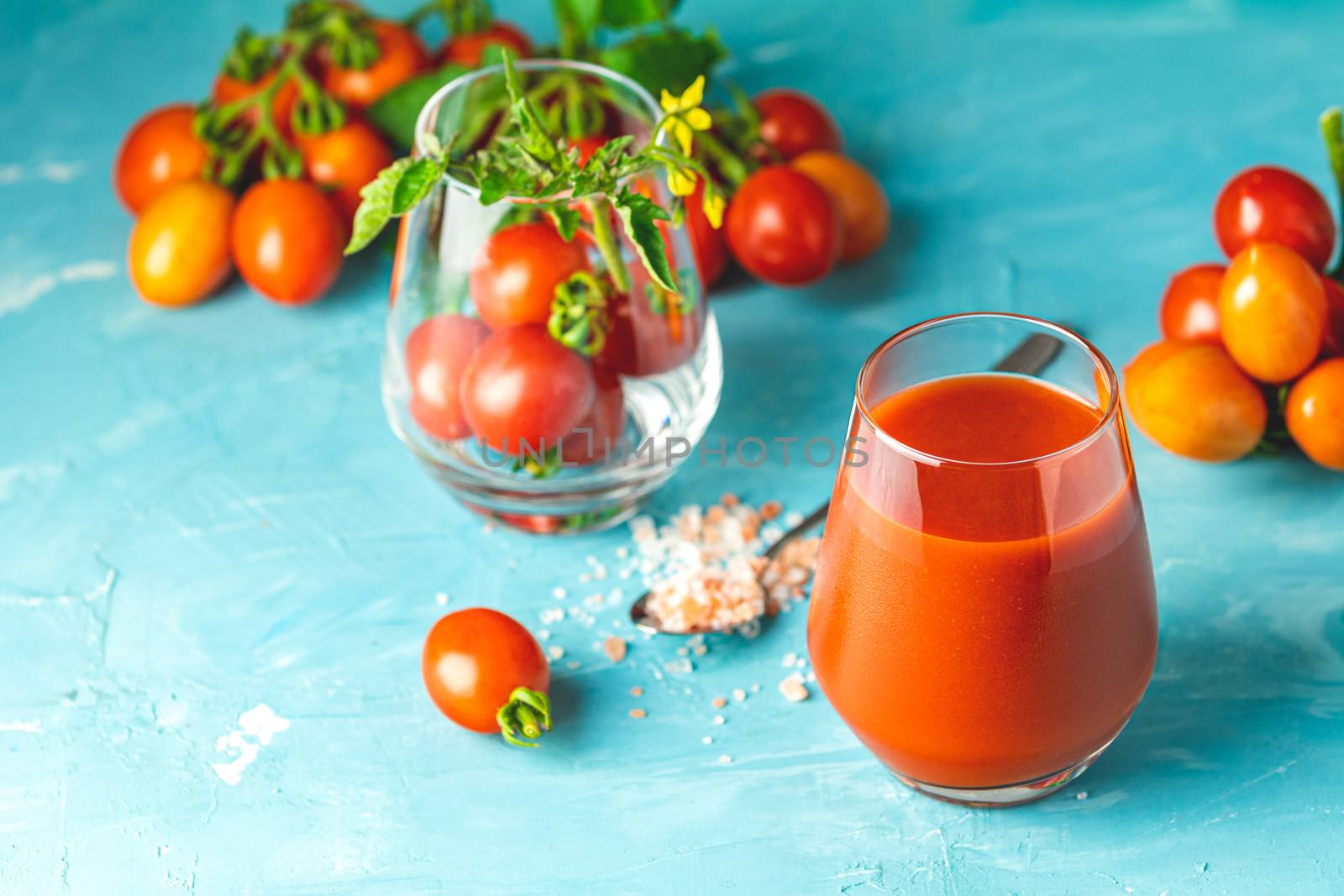 Glasses of fresh delicious jummy red tomato juice and fresh raw tomatoes with pink salt in spoon on light concrete surface. Close up. Gmo free. Natural good food.