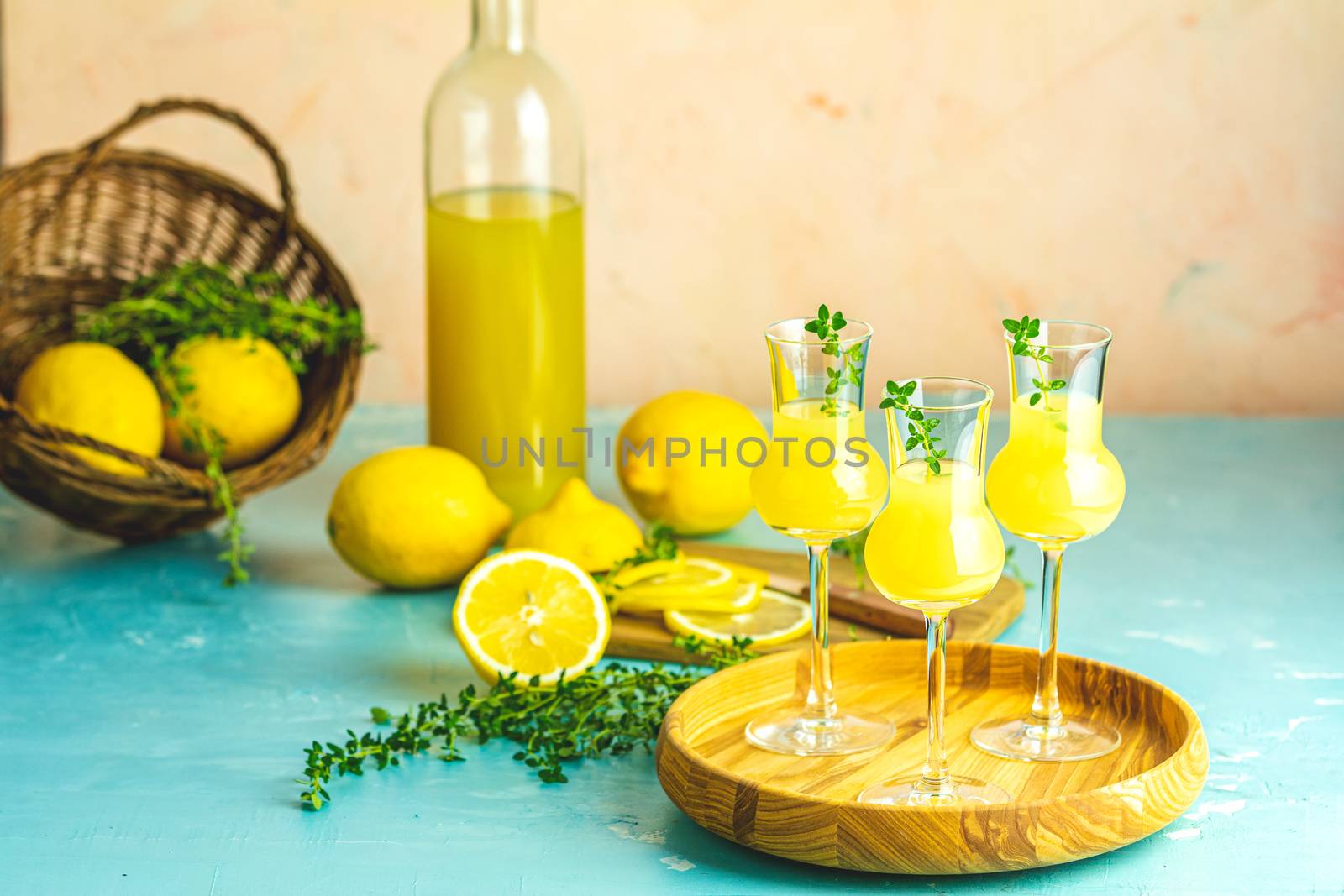 Limoncello with thyme in three grappas wineglass in wooden tray, fresh lemon in basket on light concrete table. Artistic still life on light background.