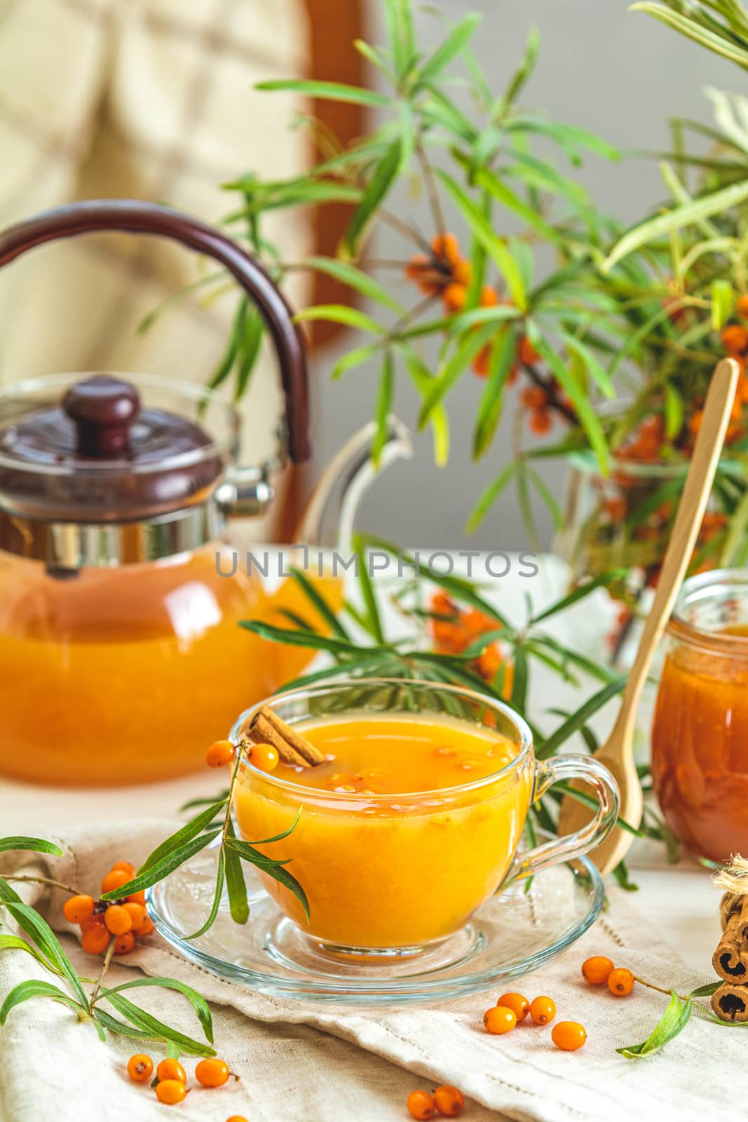 Cup and teapot of hot spicy tea with sea buckthorn, jam in the glass jar, branches of fresh berries on light wooden table surface in the rustic room