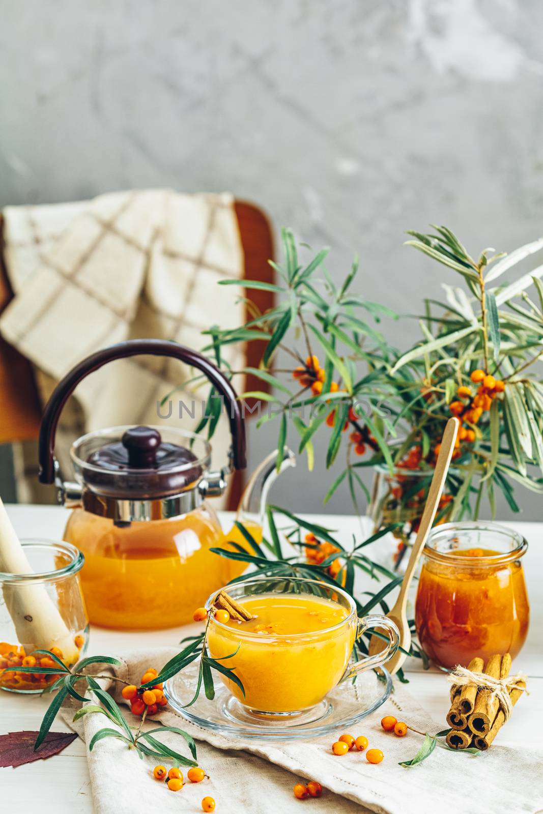 Cup and teapot of hot spicy tea with sea buckthorn, jam in the glass jar, branches of fresh berries on light woden table surface in the rustic room
