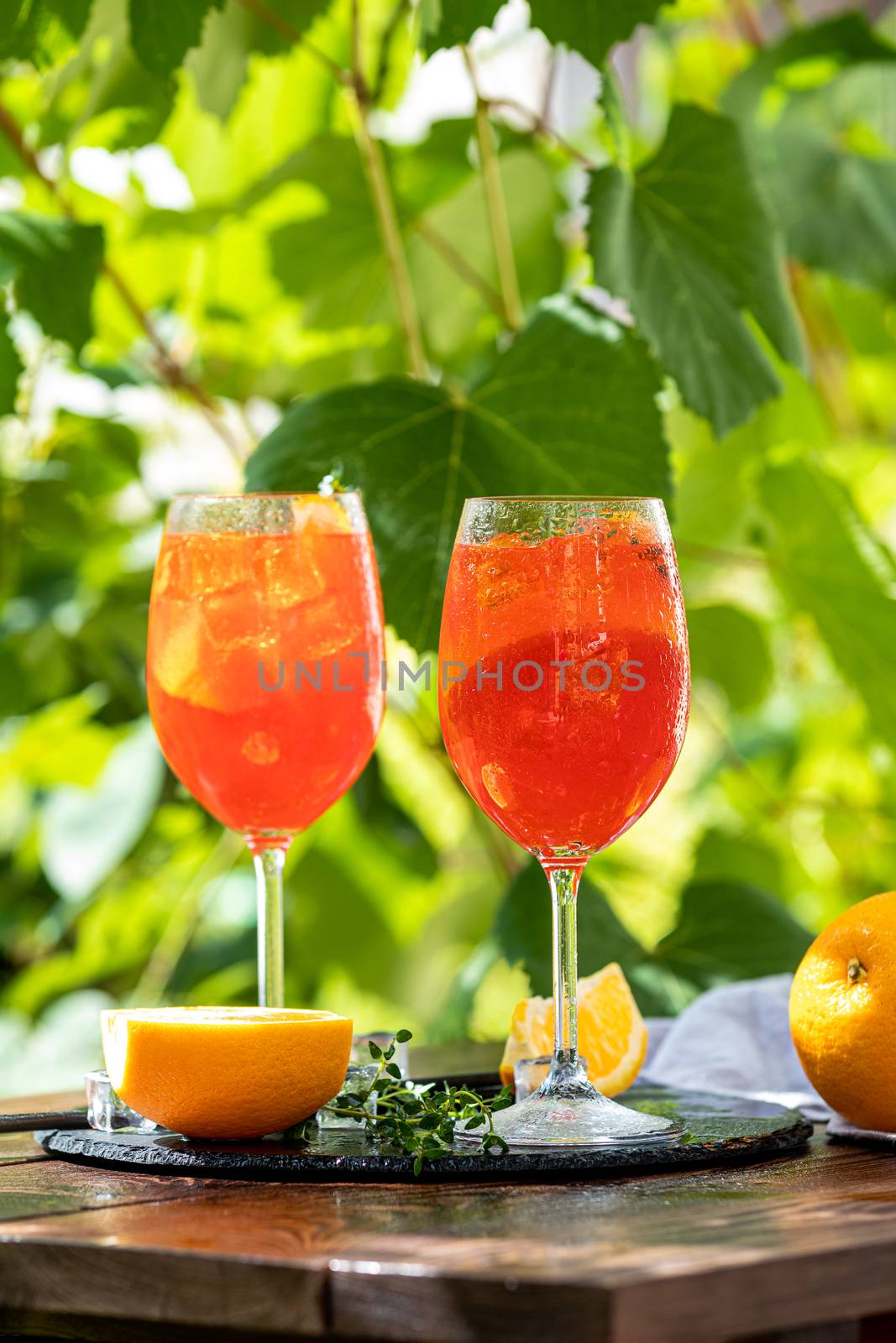 Two Aperol spritz cocktail in big wine glass with oranges, summer Italian fresh alcohol cold drink. Sunny garden with vineyard background	