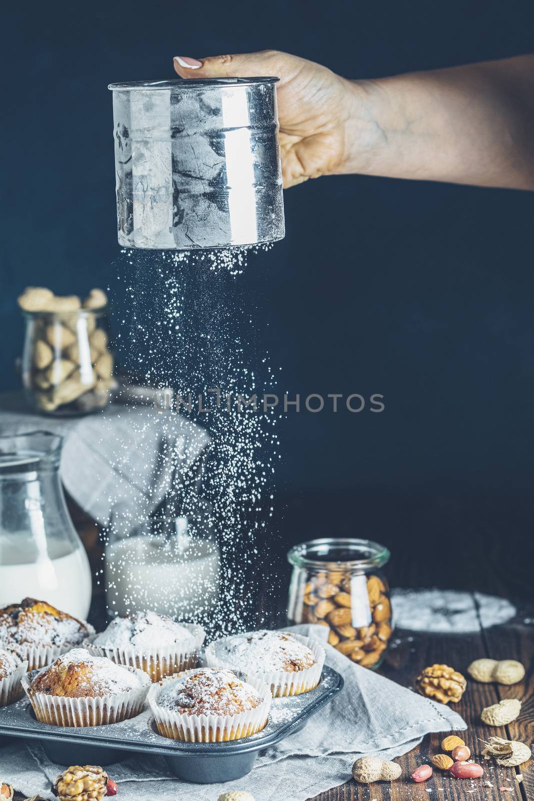 Aged female hand sifting powdered sugar by sieve over vanilla caramel muffins in paper cups in bakeware. Sifting powdered sugar on delicious cupcake with raisins, almonds and nuts in kitchen