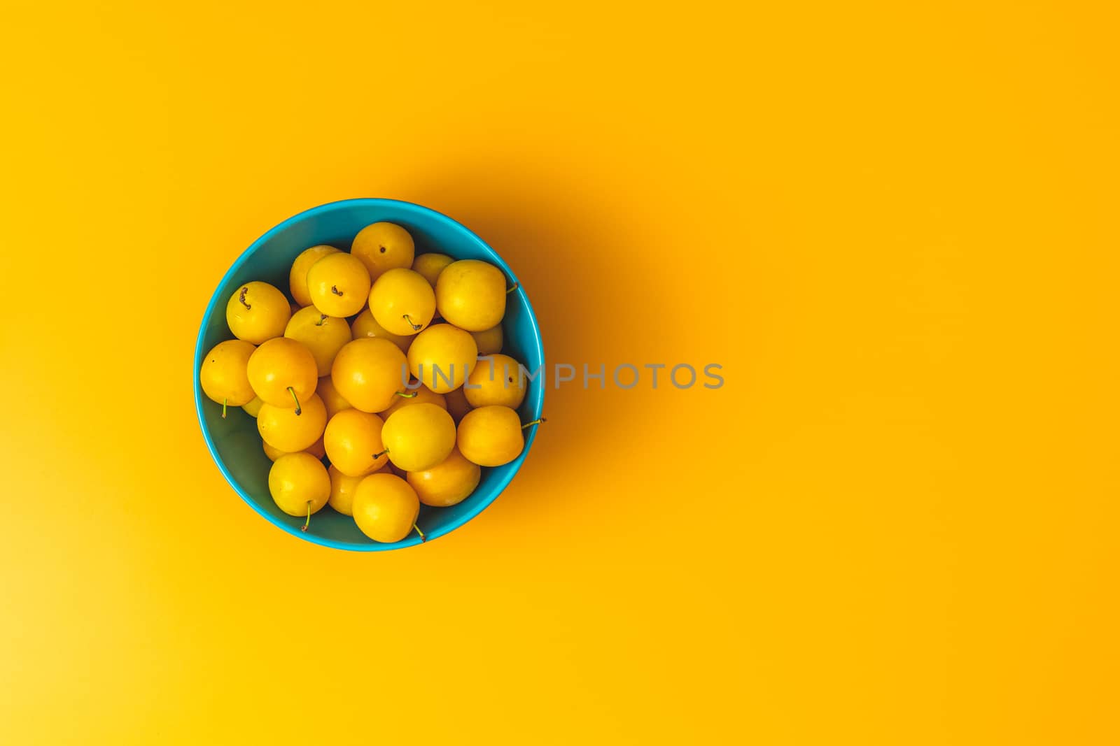 Creative summer pattern made of fresh yellow cherry plums in blue bowl on pastel yellow background. Fruit minimal concept. Flat lay.
