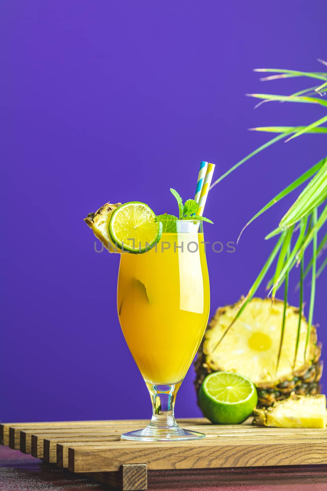 Cold cocktail with squeezed pineapple juice, lime and mint in tall glass on violet background. Summer drinks and alcoholic cocktails.  