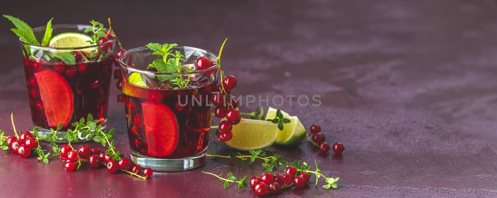 Panoramic view to two glasses of cold red cocktail with currant, lime, mint, thyme and ice in glass on dark red concrete surface table. Summer drinks and alcoholic cocktails