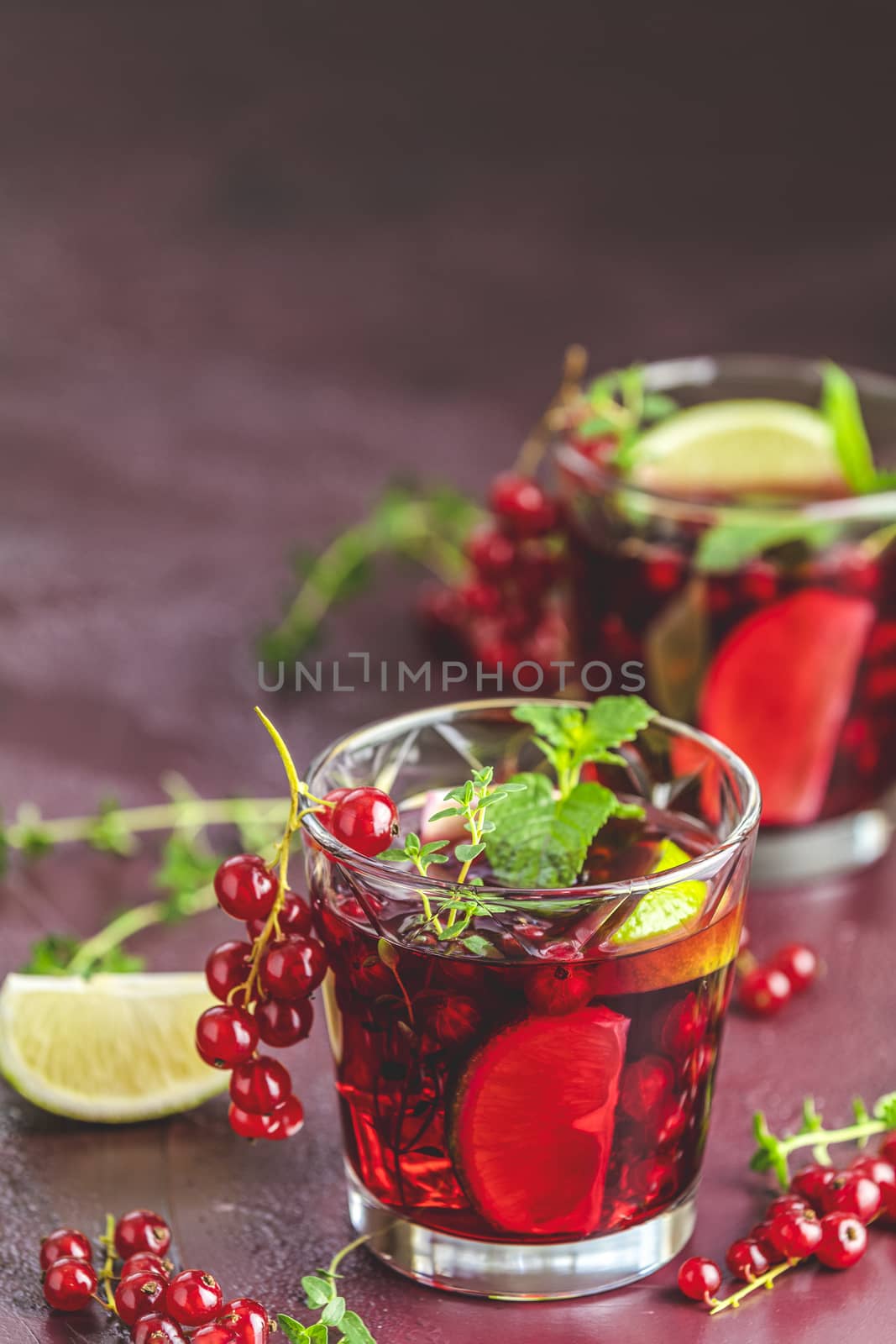 Two glasses of cold red cocktail with currant, lime, mint, thyme and ice in glass on dark red concrete surface table. Summer drinks and alcoholic cocktails. Alcoholic cocktail Currant mojito.