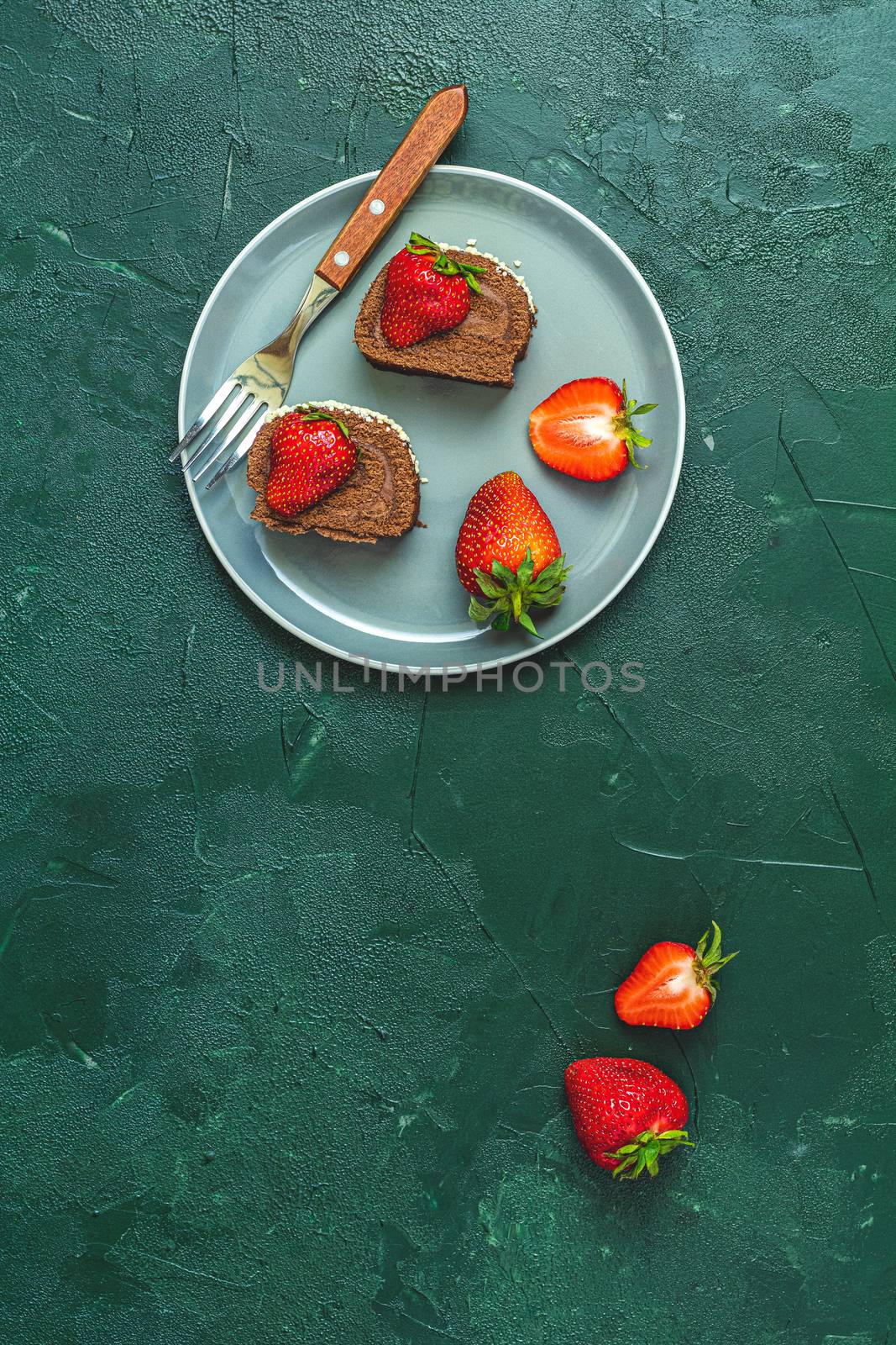 Chocolate rollcake with fresh strawberries in ceramic plate, jasmine and white peonies on dark green concrete surface table. Top view, flat lay, copy space for you text.