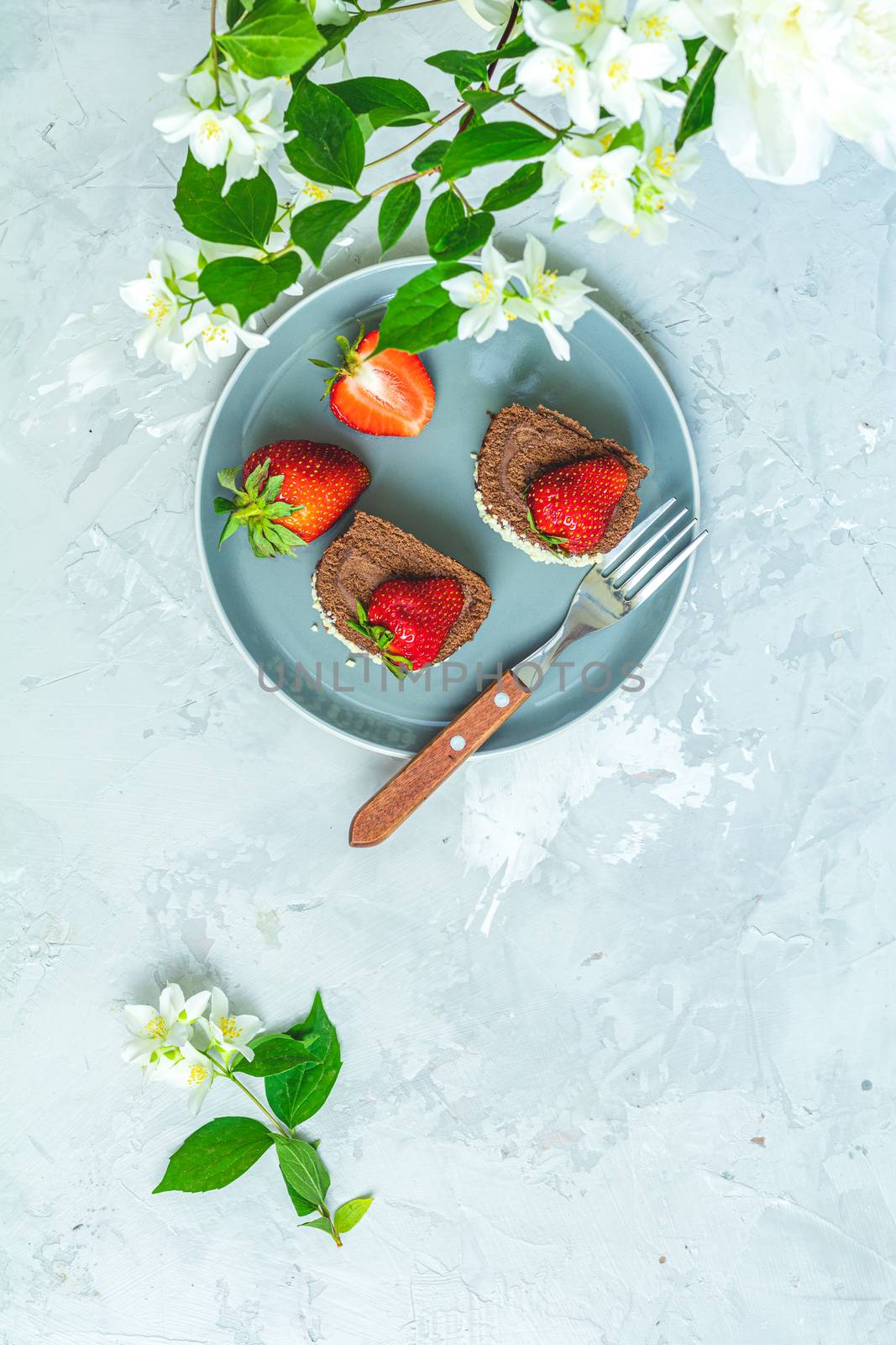 Chocolate rollcake with fresh strawberries on ceramic plate, jasmine and white peonies on light gray concrete surface table. Top view, flat lay, copy space for you text