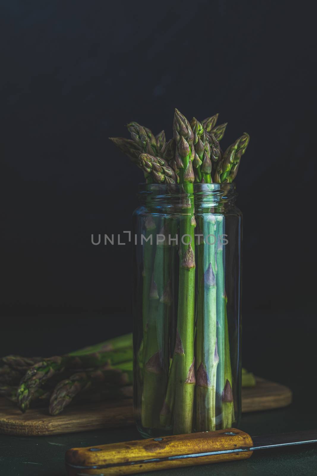 Green asparagus in glass jar standing on table at dark wall background. Fresh asparagus store. Spring seasonal vegetables. Copy space 