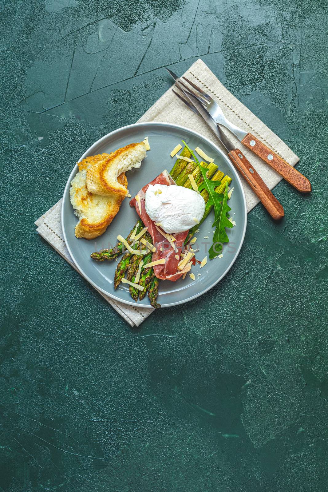 Green asparagus and boiled poached egg with hamon (jamon) and chopped parmigiano (parmesan) on a dark green plate over concrete background