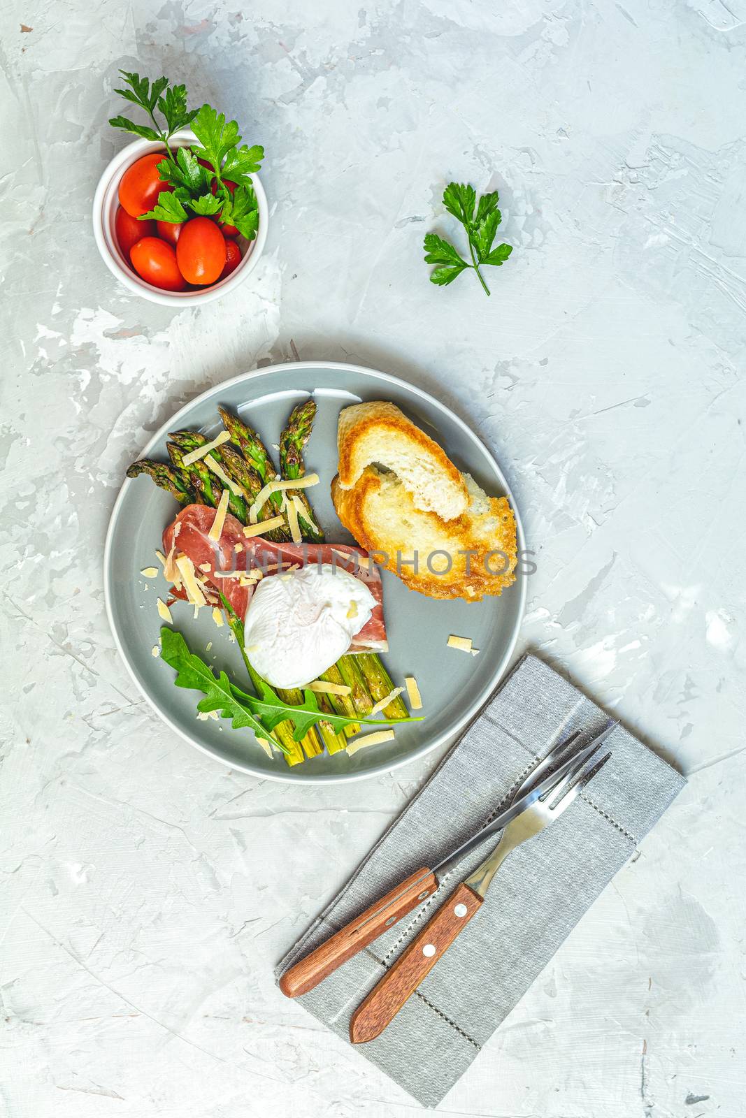 Green asparagus and boiled poached egg with hamon (jamon) and chopped parmigiano (parmesan) on a light gray plate over concrete background