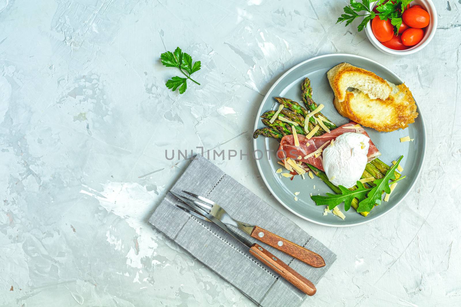 Green asparagus and boiled poached egg with hamon (jamon) and chopped parmigiano (parmesan) on a light gray plate over concrete background