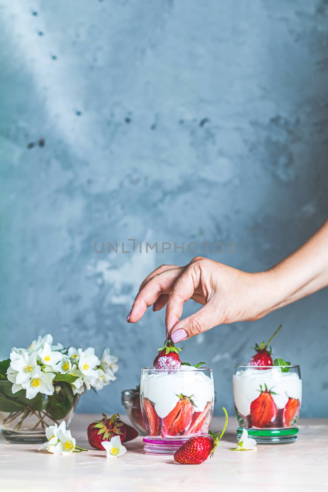 Woman hand holding cream strawberry. Glass bowl of strawberries with whipped cream and mint. Jasmine flowers. Concrete surface, copy space  for you text.