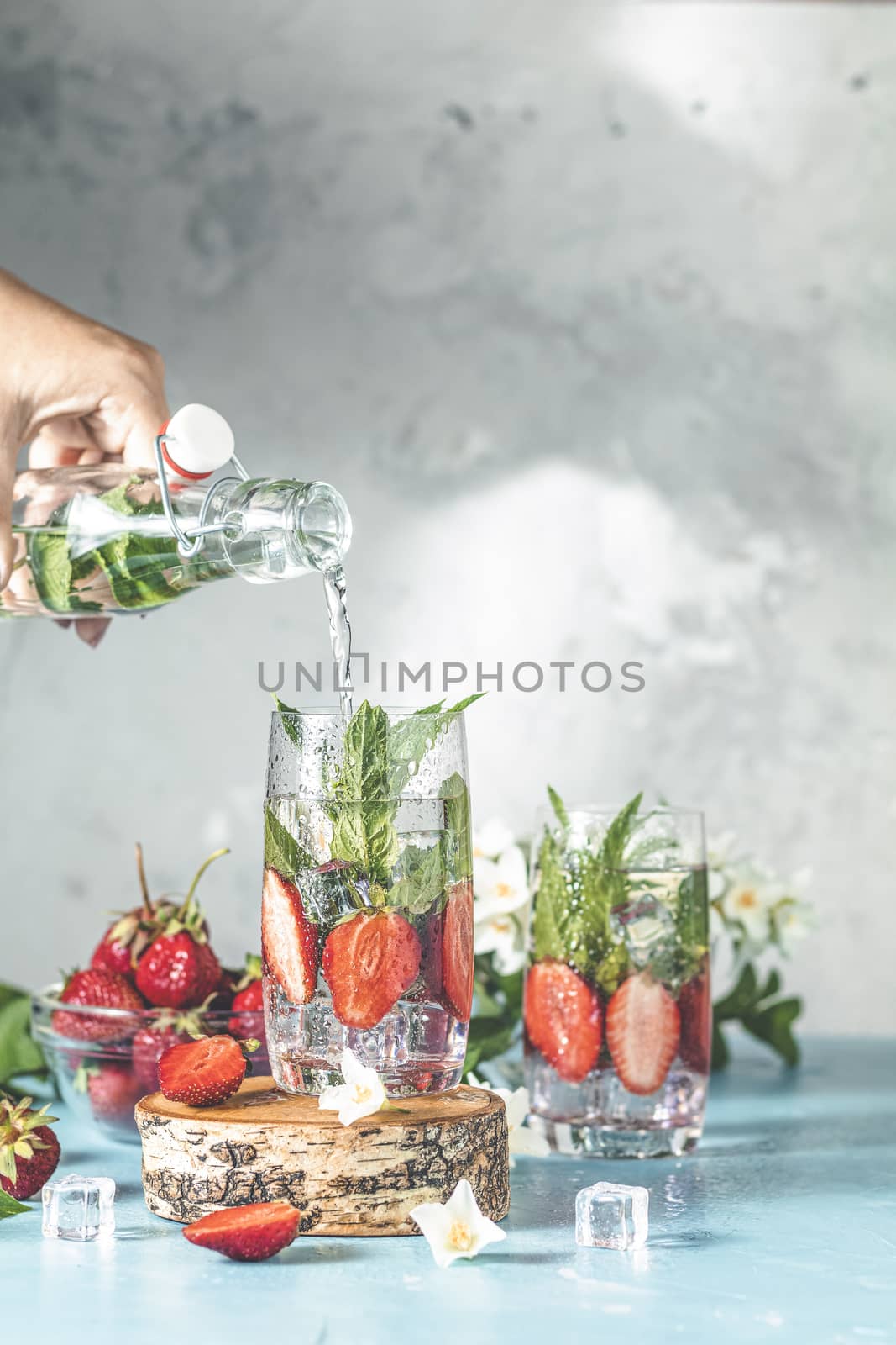 Women hand is pouring detox water from glass bottle to glass with strawberries, ice and mint. Summer fresh Mint soda cocktail, selective focus.