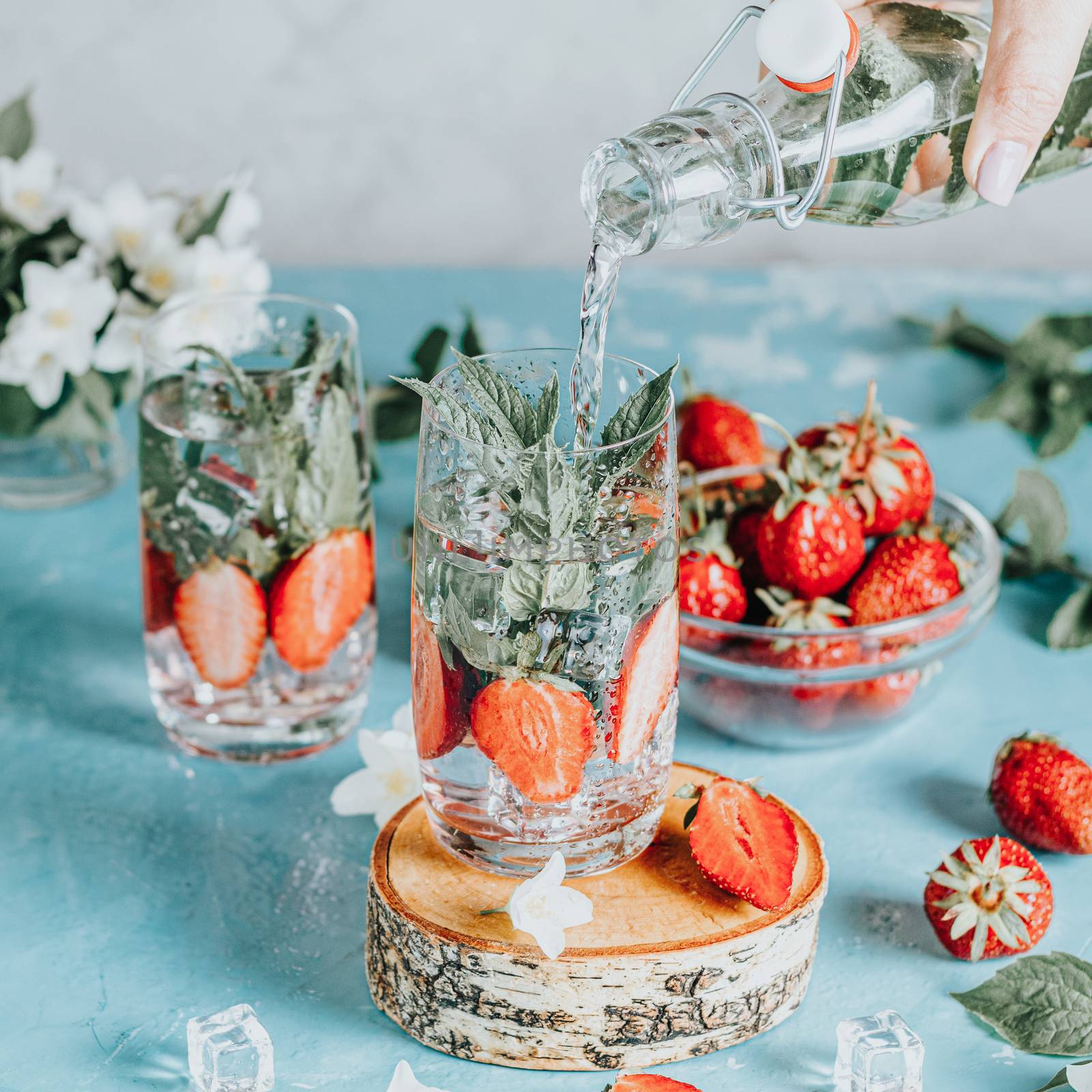 Woman pouring lemonade from bottle into glass. Detox infused water with strawberry and mint in highball glasses on blue concrete table background, copy space. Cold summer drink. Mineral water.