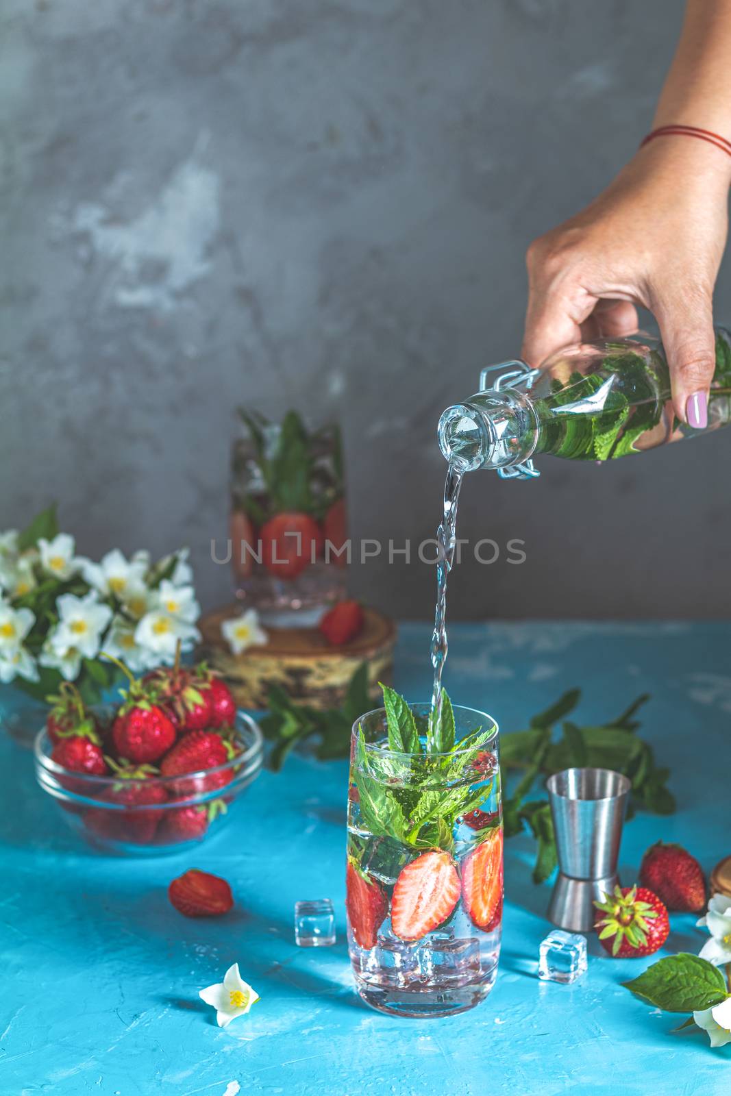 Woman pouring lemonade from bottle into glass. Detox infused water with strawberry and mint in highball glasses on blue concrete table background, copy space. Cold summer drink. Mineral water.