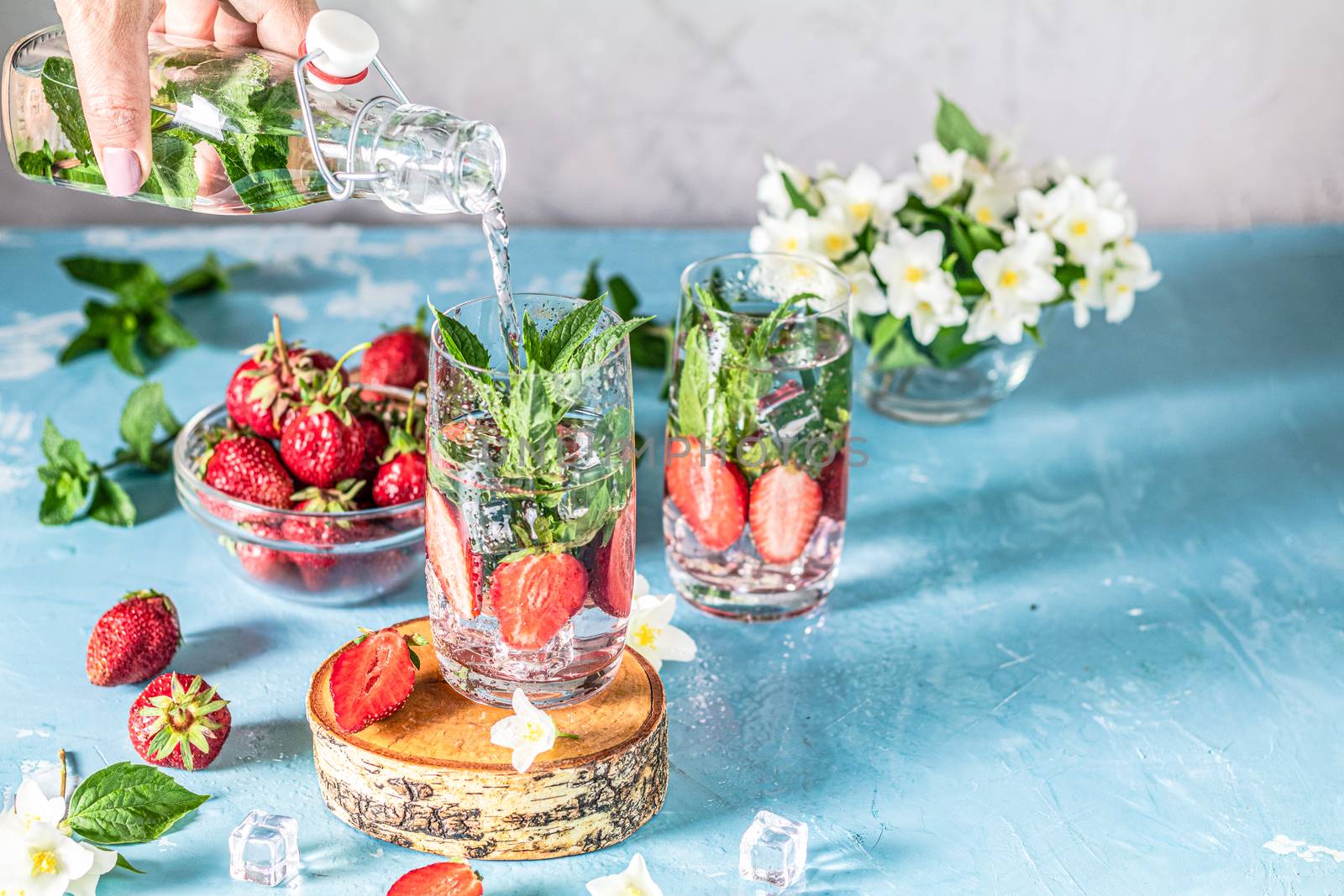 Woman pouring lemonade from bottle into glass. Detox infused water with strawberry and mint in highball glasses on blue concrete table background, copy space. Cold summer drink. Mineral water.