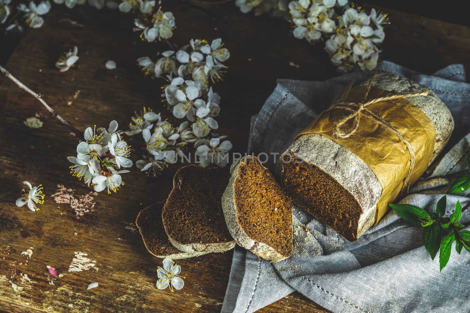 Sliced freshly baked rye handmade breads on old wooden table with linen napkin and apricot tree blossom branch. Dark rustic style.