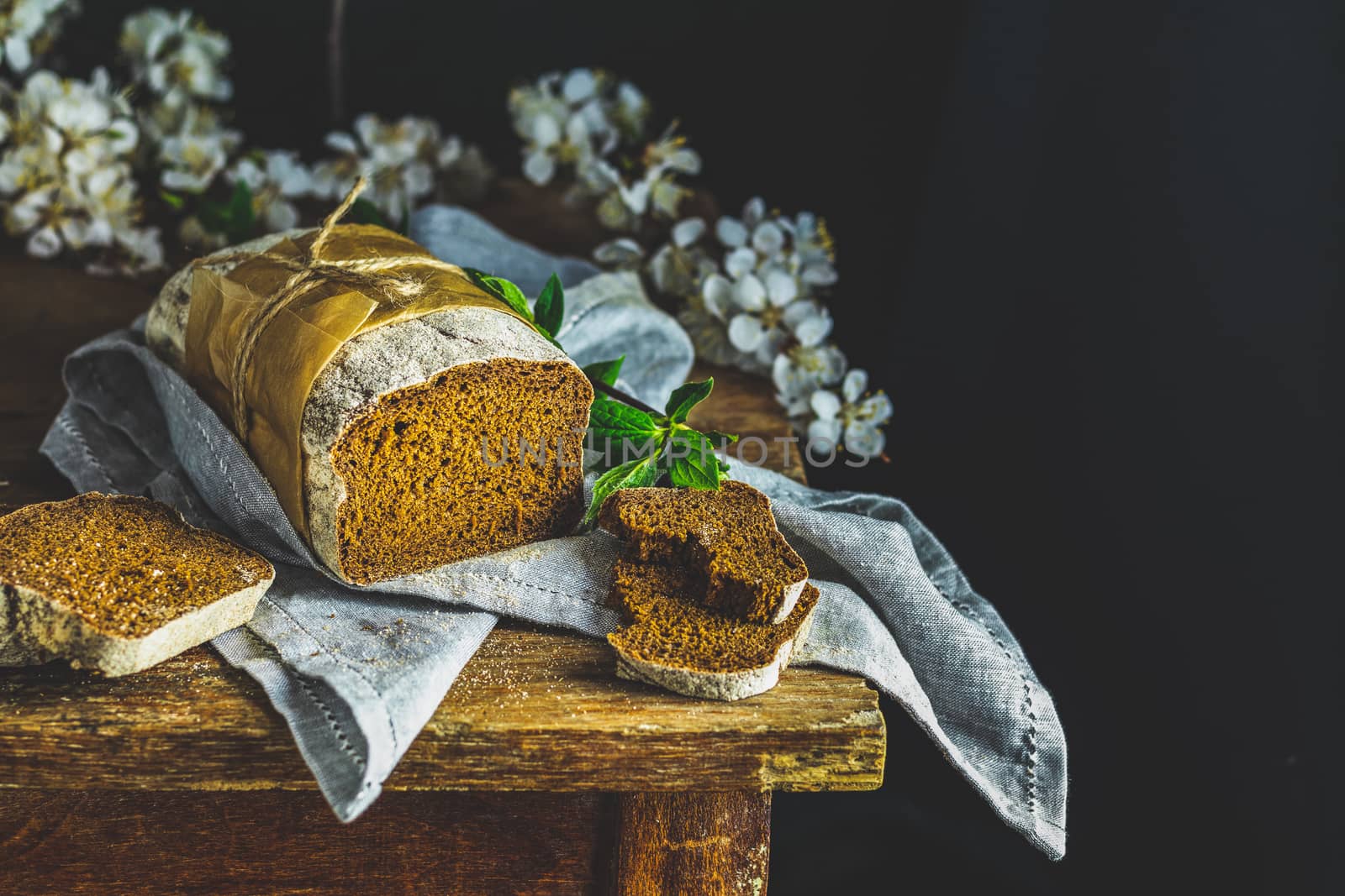Sliced freshly baked rye handmade breads on old wooden table with linen napkin and apricot tree blossom branch. Dark rustic style.