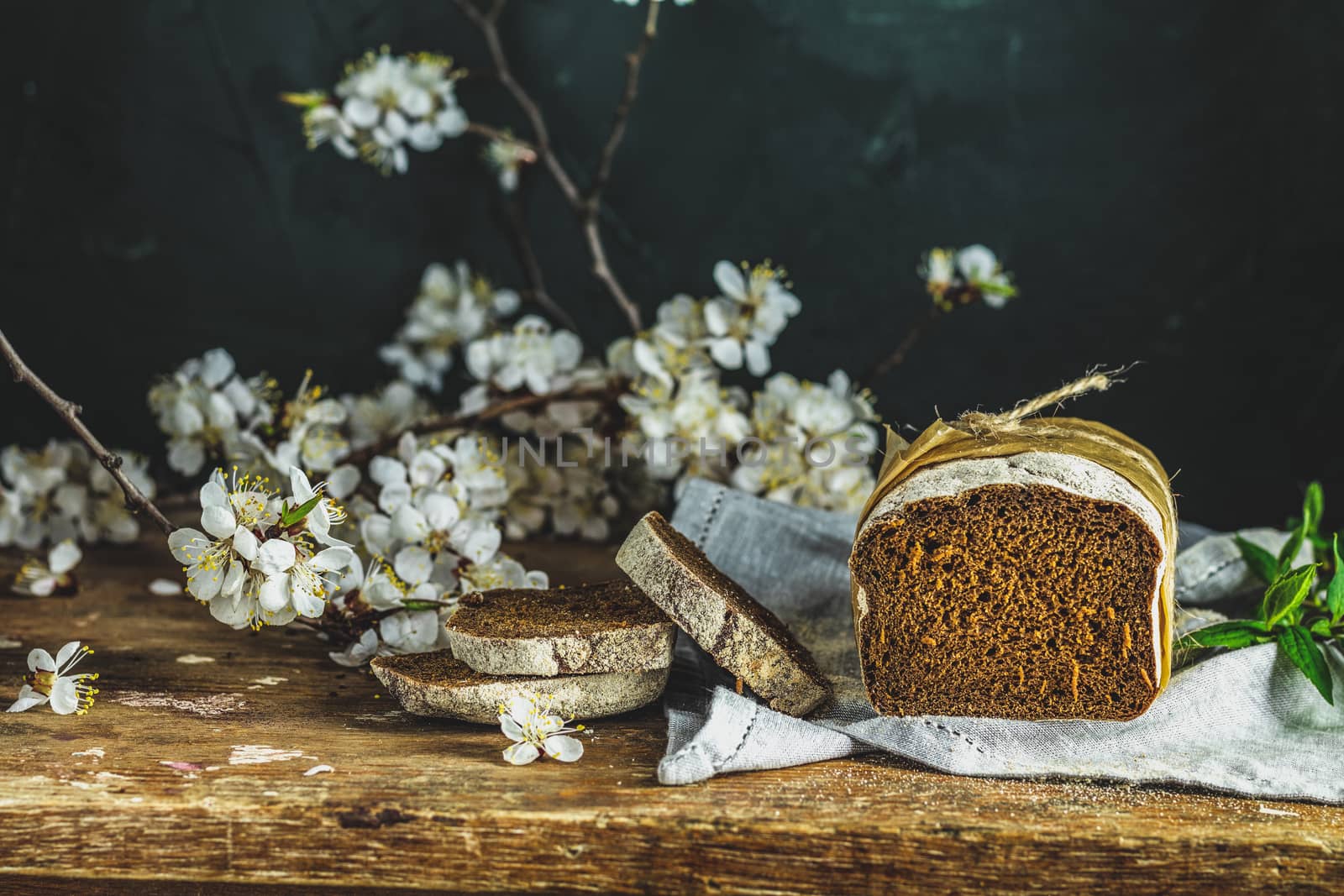 Sliced freshly baked rye handmade breads on old wooden table with linen napkin and apricot tree blossom branch. Dark rustic style.