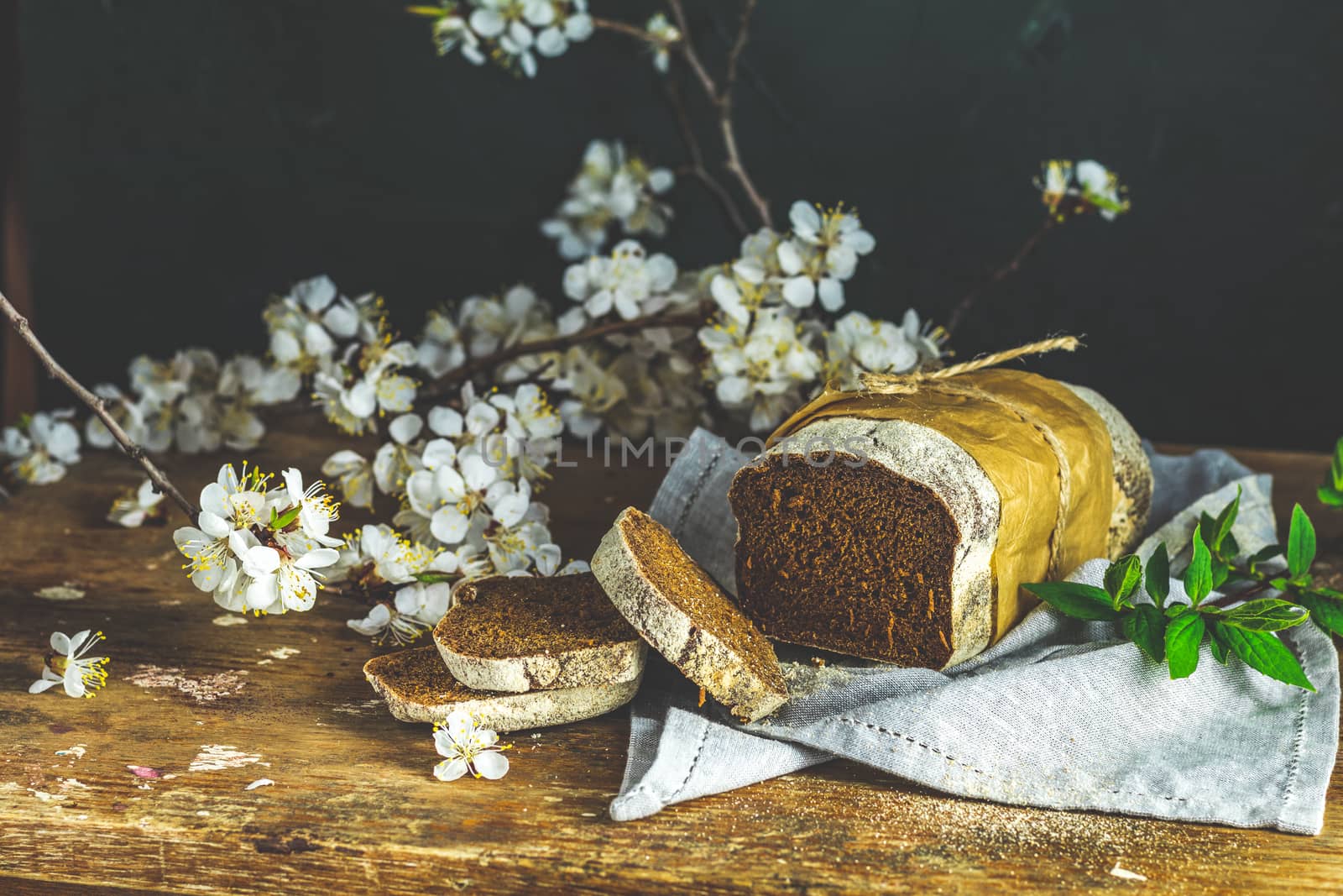 Sliced freshly baked rye handmade breads on old wooden table with linen napkin and apricot tree blossom branch. Dark rustic style.