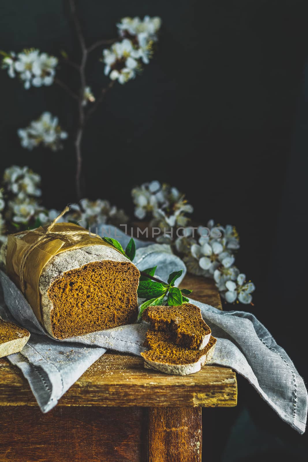 Sliced freshly baked rye handmade breads on old wooden table with linen napkin and apricot tree blossom branch. Dark rustic style.