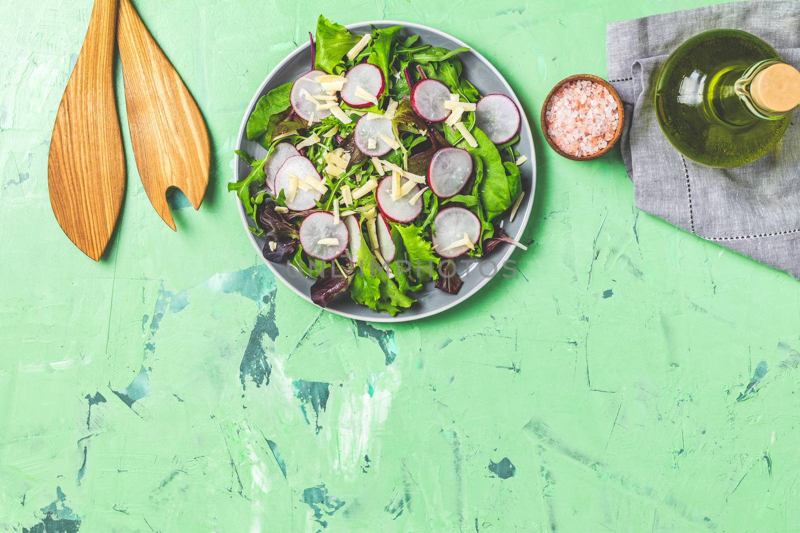 Salad with radishes, olive oil,  pink salt, empty ceramic bowl, wooden spoons for salad over green concrete concrete texture background. Top view, healthy food concept, top view, copy space for you text.