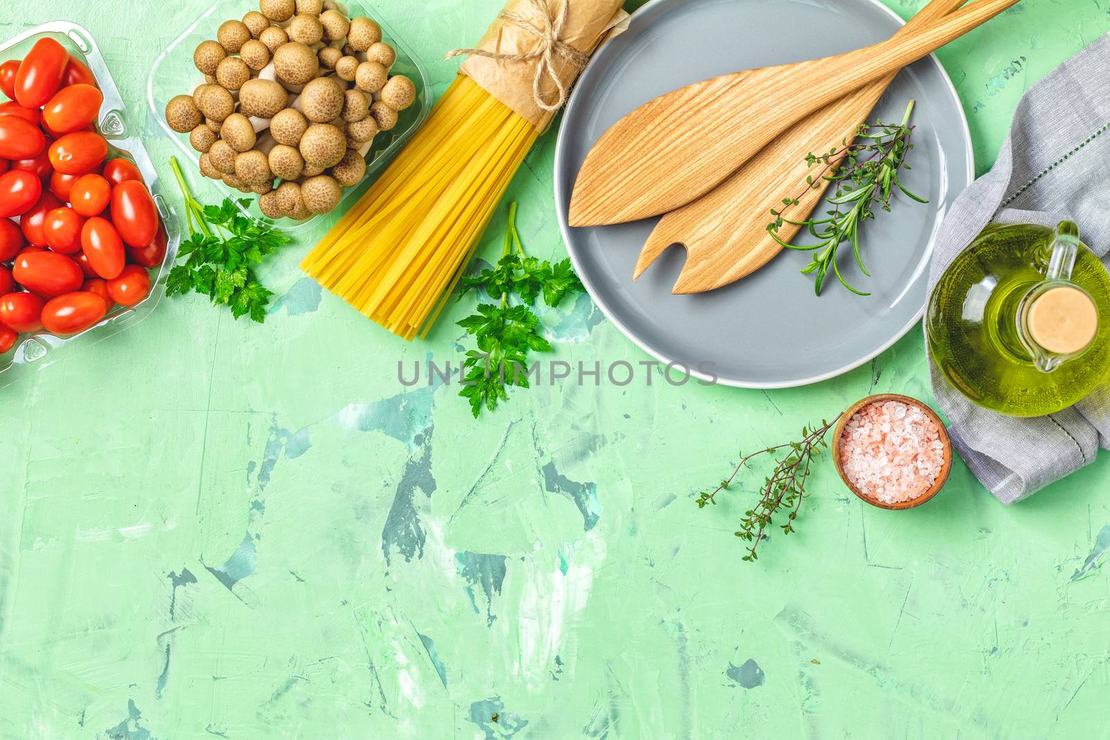 Spaghetti, shimeji mushroom, tomatoes, olive oil,  pink salt, empty ceramic bowl, wooden spoons for salad over green concrete texture background. Top view, healthy food concept, top view, copy space