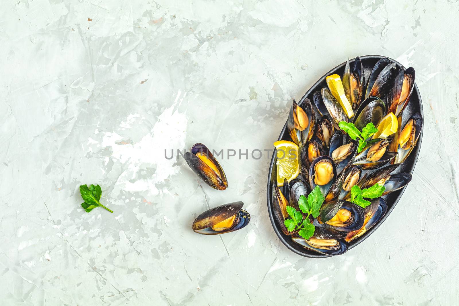 Seafood mussels with lemon and parsley in black metal plate on light gray concrete table surface, top view, copy space for you text