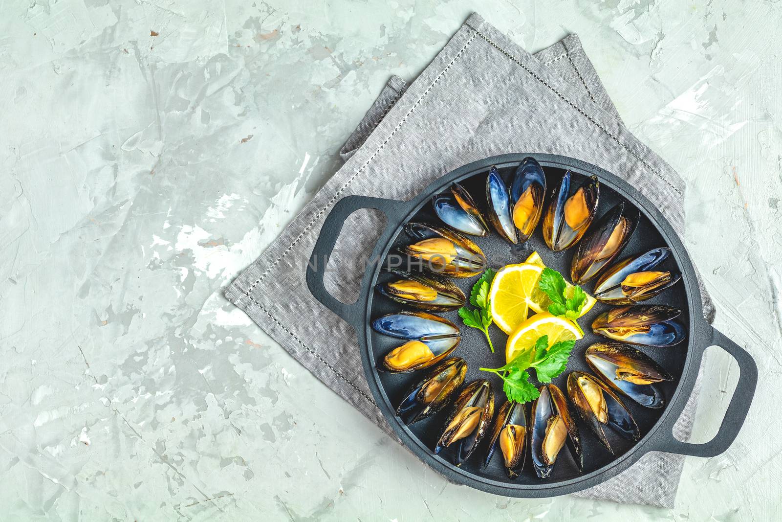Seafood mussels with lemon and parsley in black metal pan on light gray concrete table surface, top view, copy space for you text