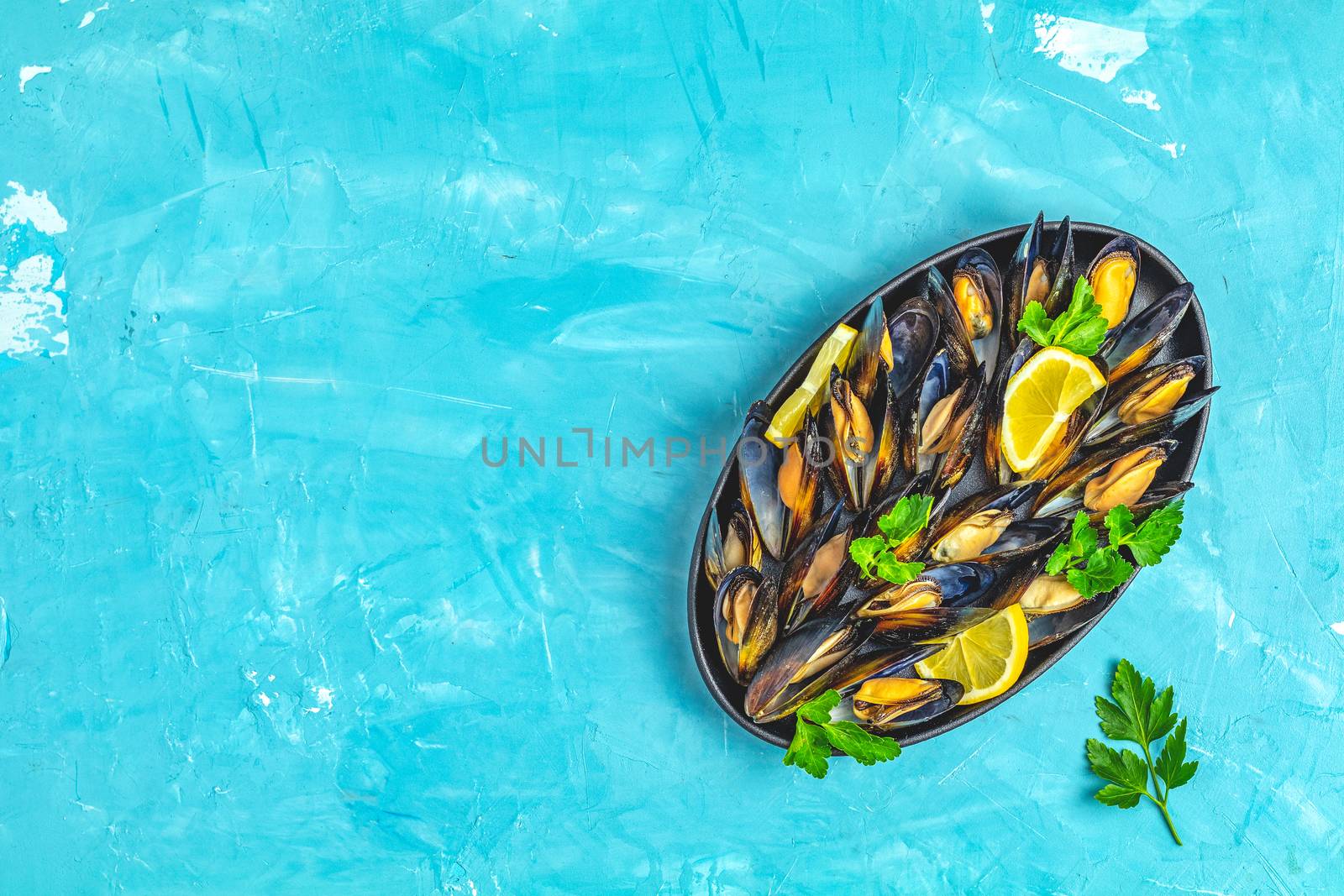 Seafood mussels with lemon and parsley in black metal plate on blue concrete table surface, top view, copy space for you text