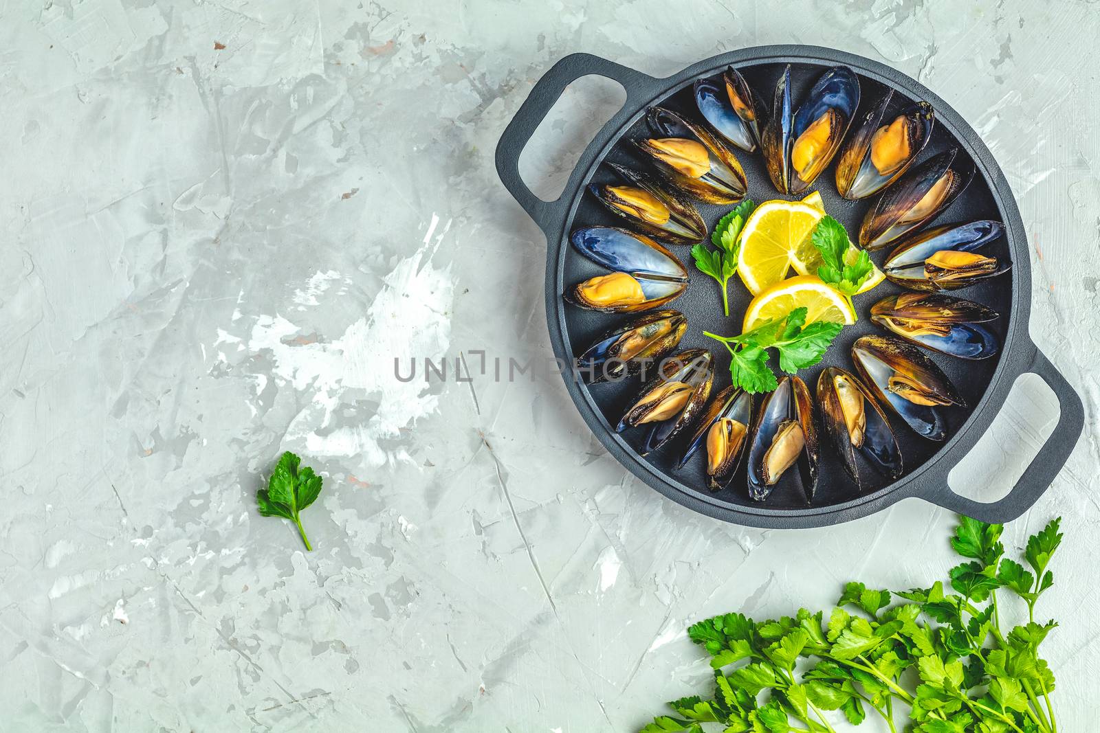 Seafood mussels with lemon and parsley in black metal pan on light gray concrete table surface, top view, copy space for you text