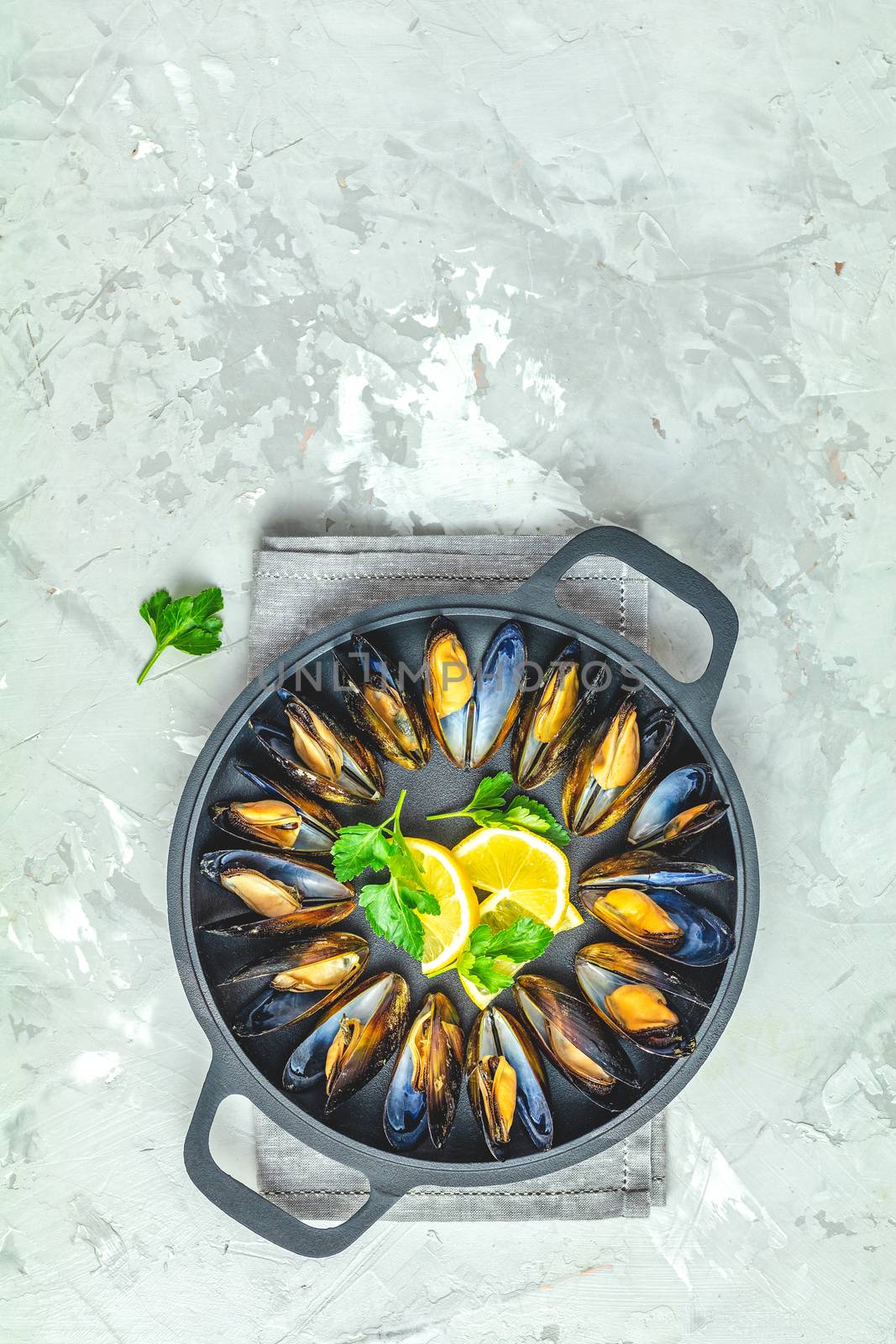 Seafood mussels with lemon and parsley in black metal pan on light gray concrete table surface, top view, copy space for you text