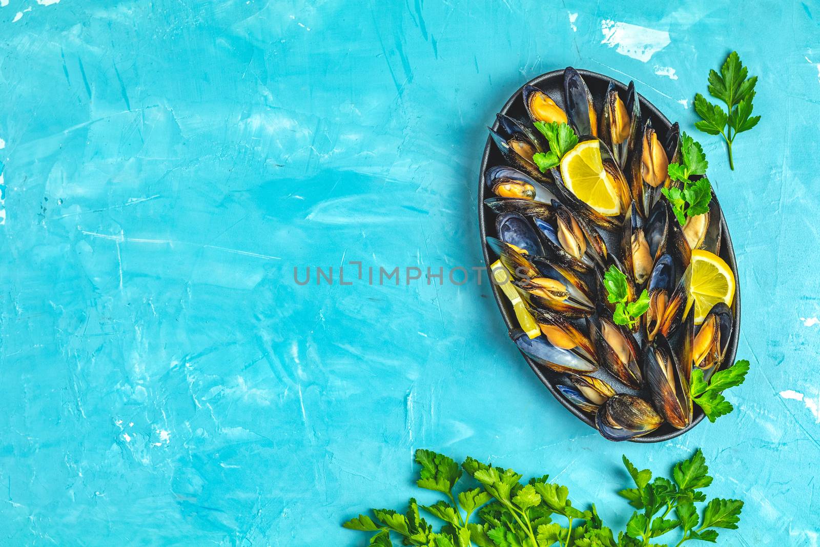 Seafood mussels with lemon and parsley in black metal plate on blue concrete table surface, top view, copy space for you text
