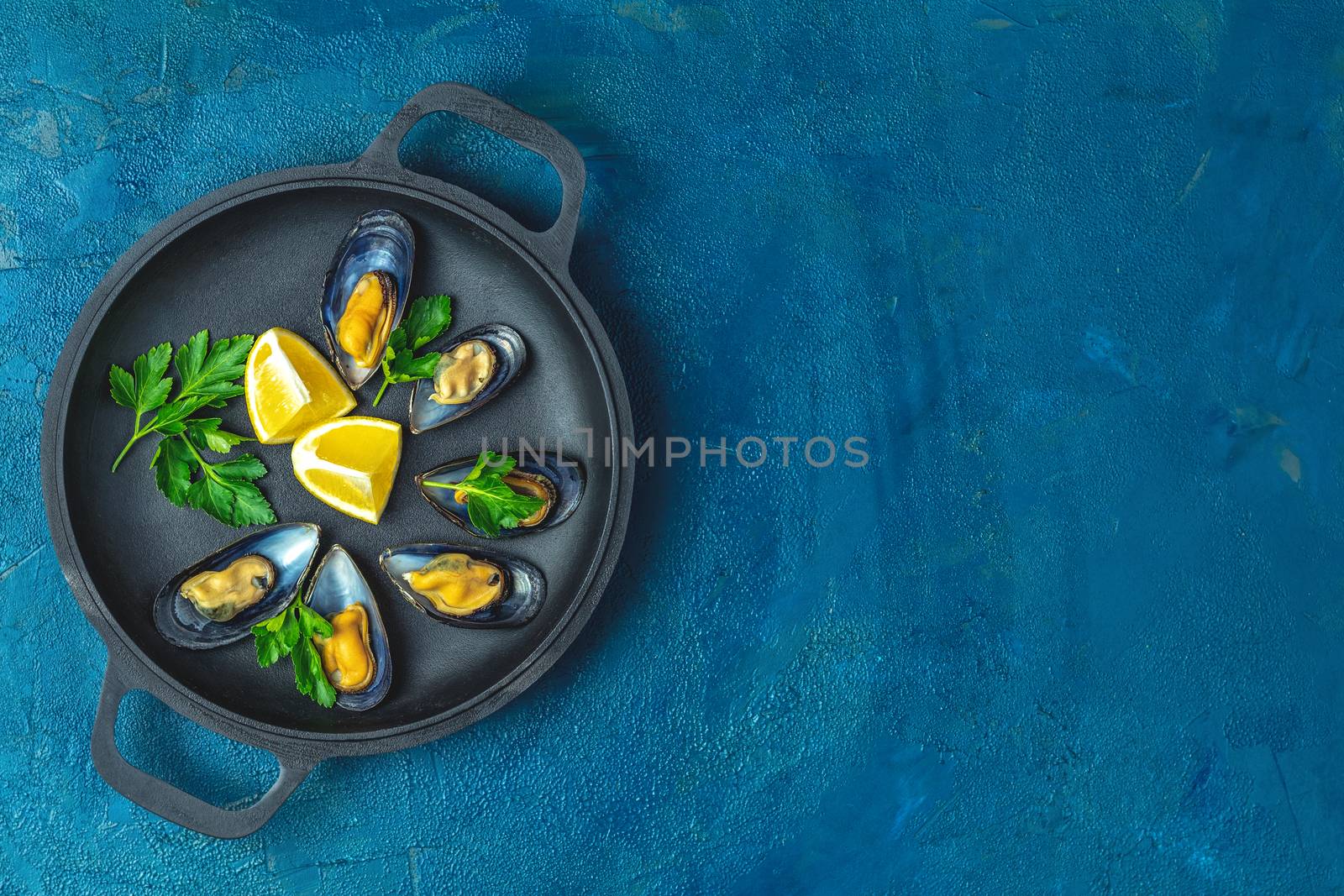Seafood mussels with lemon and parsley in black metal pan on dark blue concrete table surface, top view, copy space for you text
