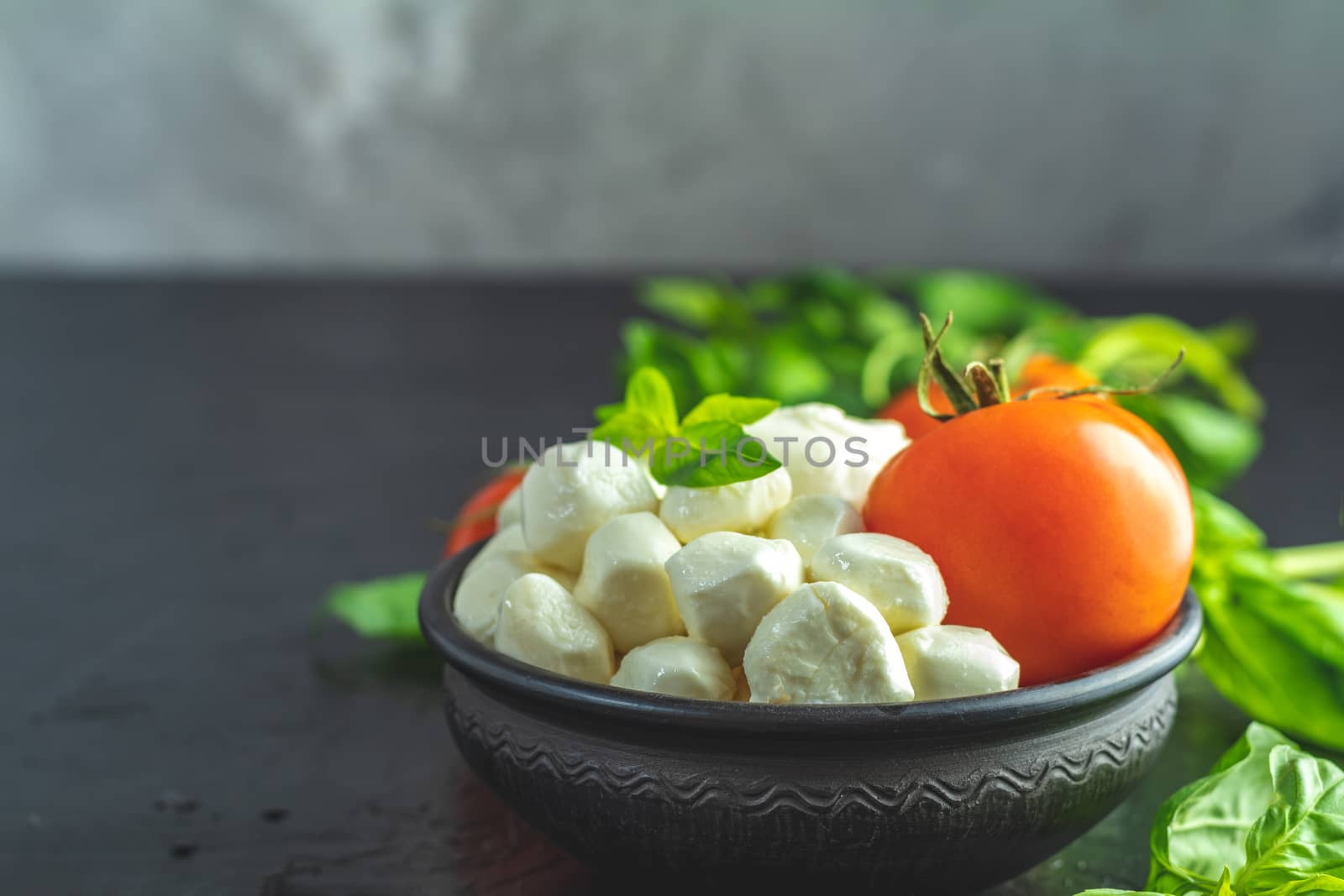 Mozzarella balls, buffalo in black ceramic plate, tomatoes and basil over dark background. With space. Rustic style. Ingredients for italian caprese salad. 